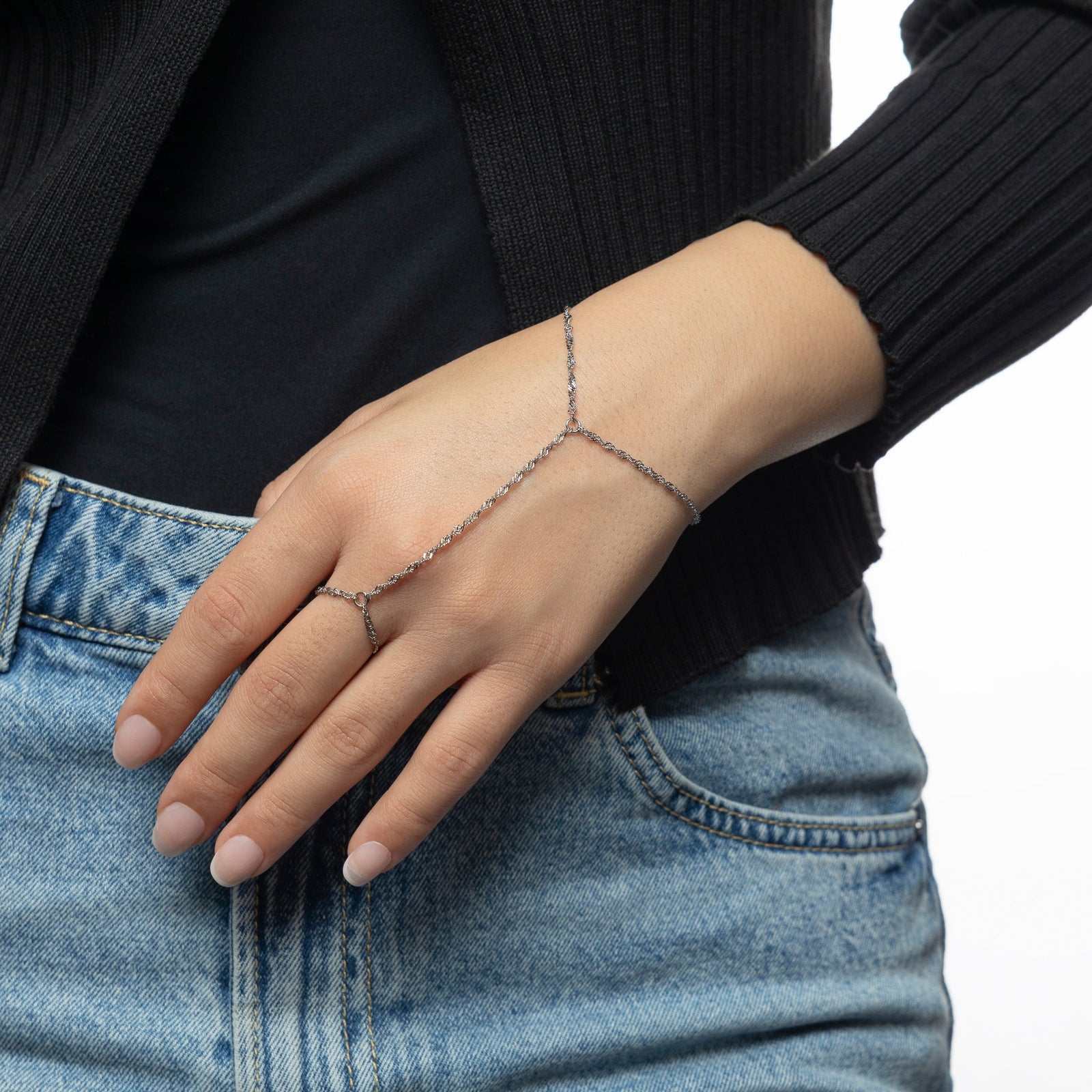 Full view of a woman’s hand in denim jeans and black top, wearing a silver twisted hand chain from wrist to middle finger.