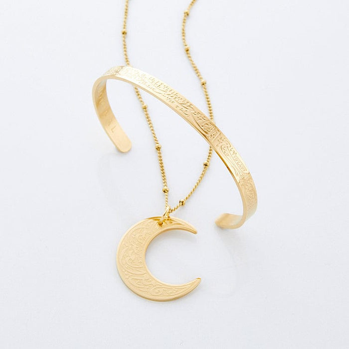 Photo of the "With Hardship Comes Ease" Crescent Necklace in Gold and "With Hardship Comes Ease" V2 Adjustable Cuff in Gold against a light background.