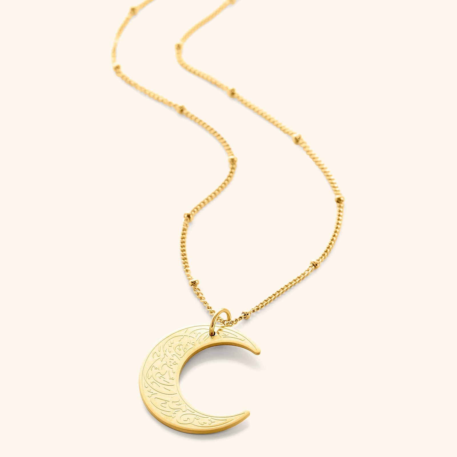 A delicate gold necklace featuring a crescent moon pendant inscribed with Arabic script. The chain is a fine gold link. The necklace is displayed on a light beige background. Given the inscription "With Hardship Comes Ease" and the crescent moon, which holds significance in Islamic culture, it's plausible that the script is related to Islamic teachings and the necklace carries religious or inspirational meaning for the wearer.