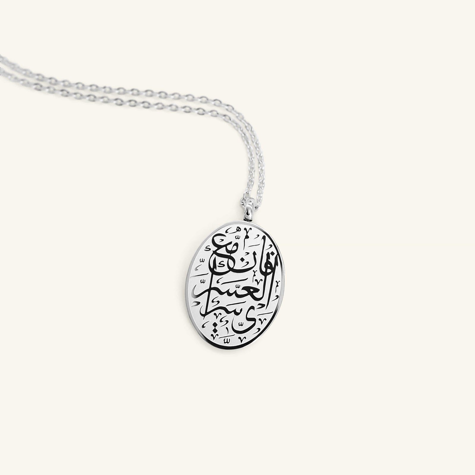 Flat lay of the silver necklace that features an oval-shaped piece engraved with an inscription that means "with hardship comes ease" (94:5)
