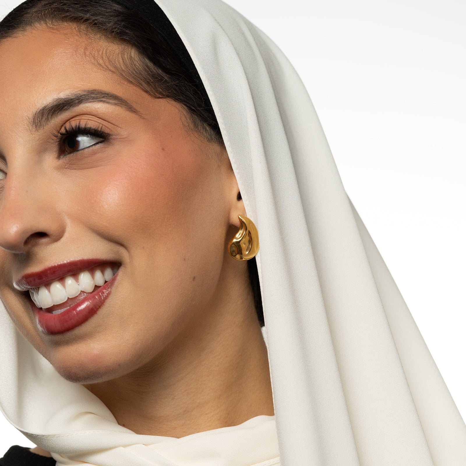 Woman wearing a white headscarf and gold vintage drople earring on a plain background