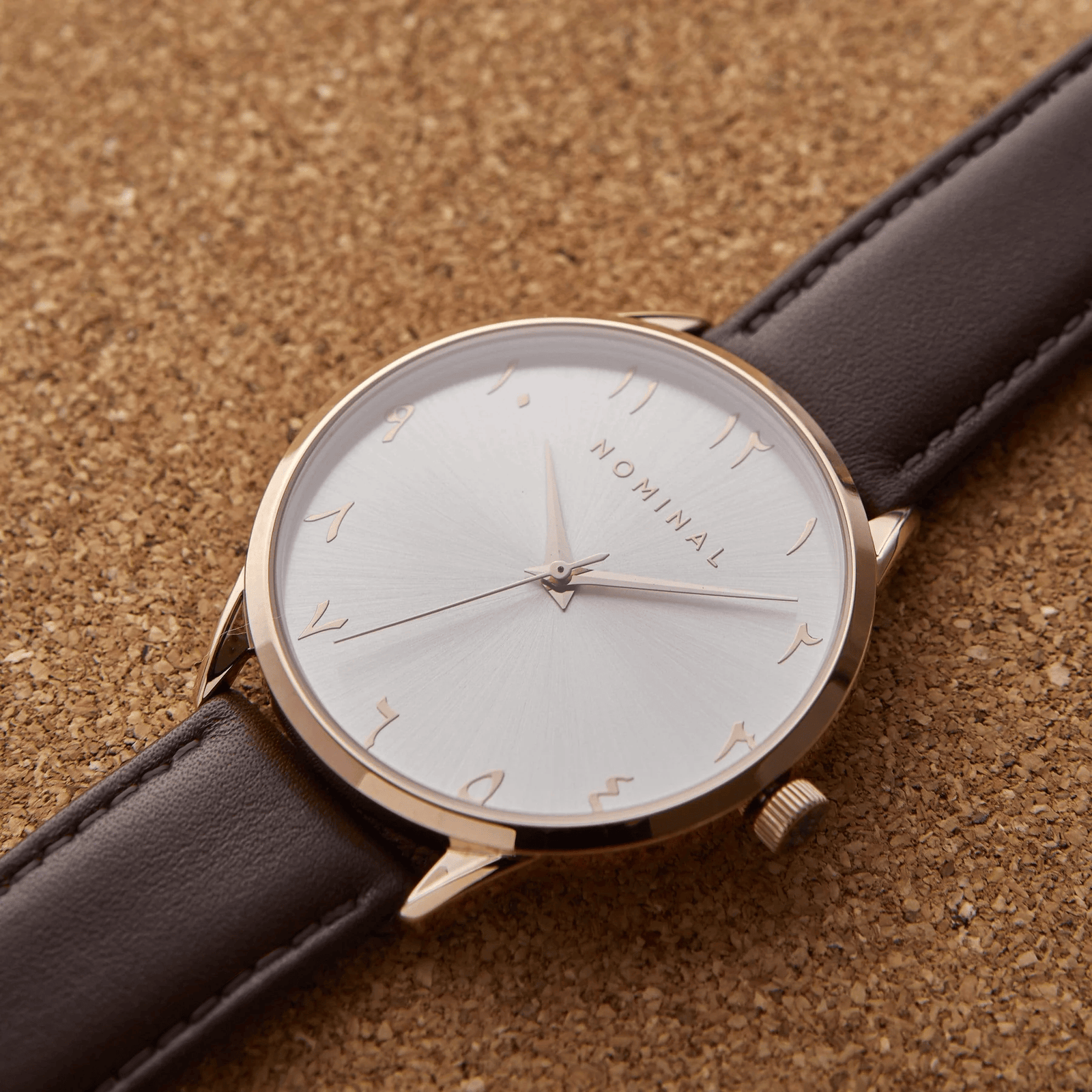 Photo of a watch with round white sunburst dial on a brown Italian Leather strap. The watch features Rose Gold Case, Rose gold numbers, NOMINAL logo, and pin.