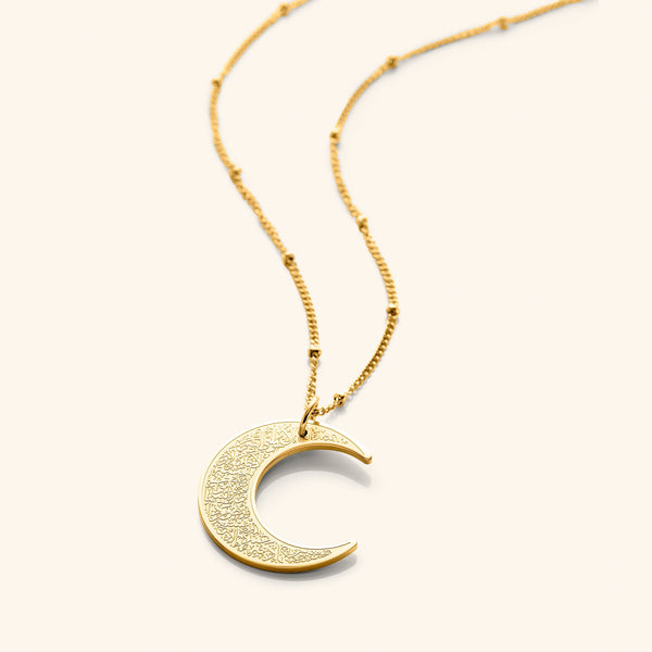 A close-up of the Ayatul Kursi Crescent Necklace in 18K gold plating, featuring a crescent-shaped pendant intricately engraved with Arabic calligraphy, suspended from a delicate cable chain.