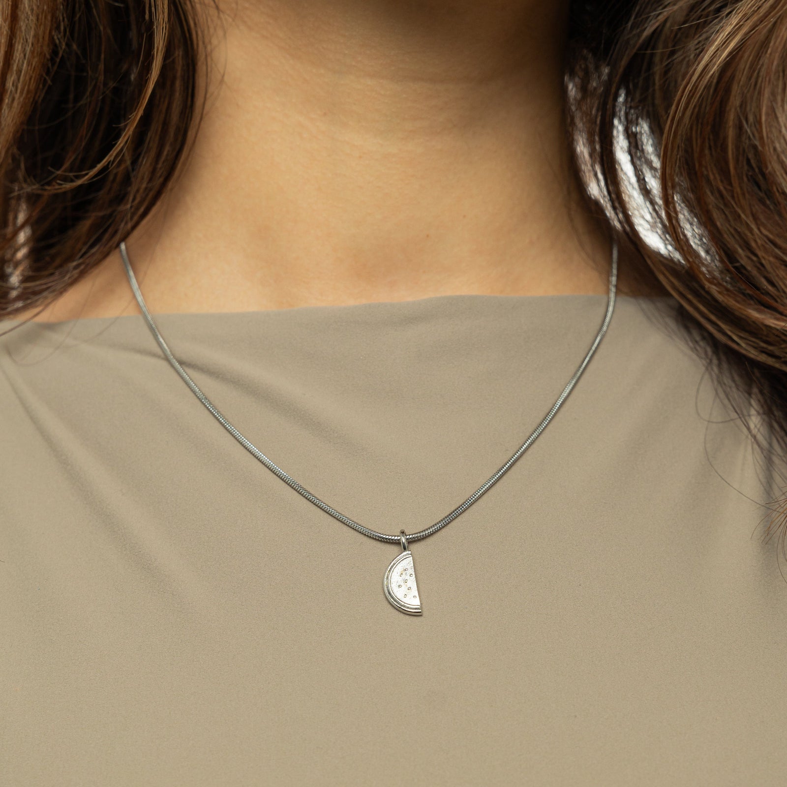 A close-up shot of a person wearing a delicate silver necklace with a small, detailed pendant shaped like a slice of a watermelon. The pendant hangs on a thin silver chain that rests against a light olive-green top. The person has light brown, wavy hair and light-toned skin. Only the lower part of their face, neck, and upper chest are visible.