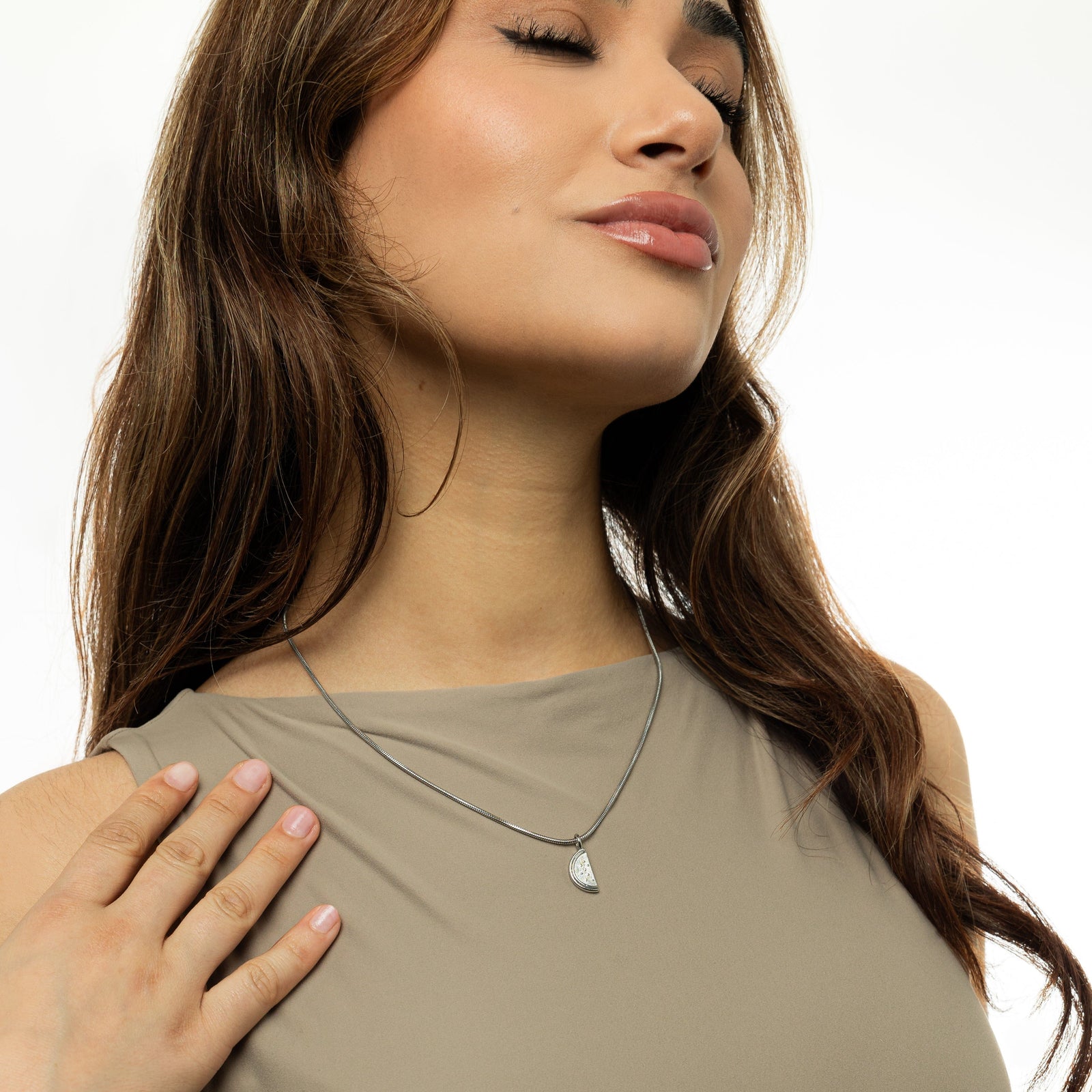 A close-up shot of a person wearing a delicate silver necklace with a small, detailed pendant shaped like a slice of a watermelon. The pendant hangs on a thin silver chain that rests against a light olive-green top. The person has light brown, wavy hair and light-toned skin. Only the lower part of their face, neck, and upper chest are visible.