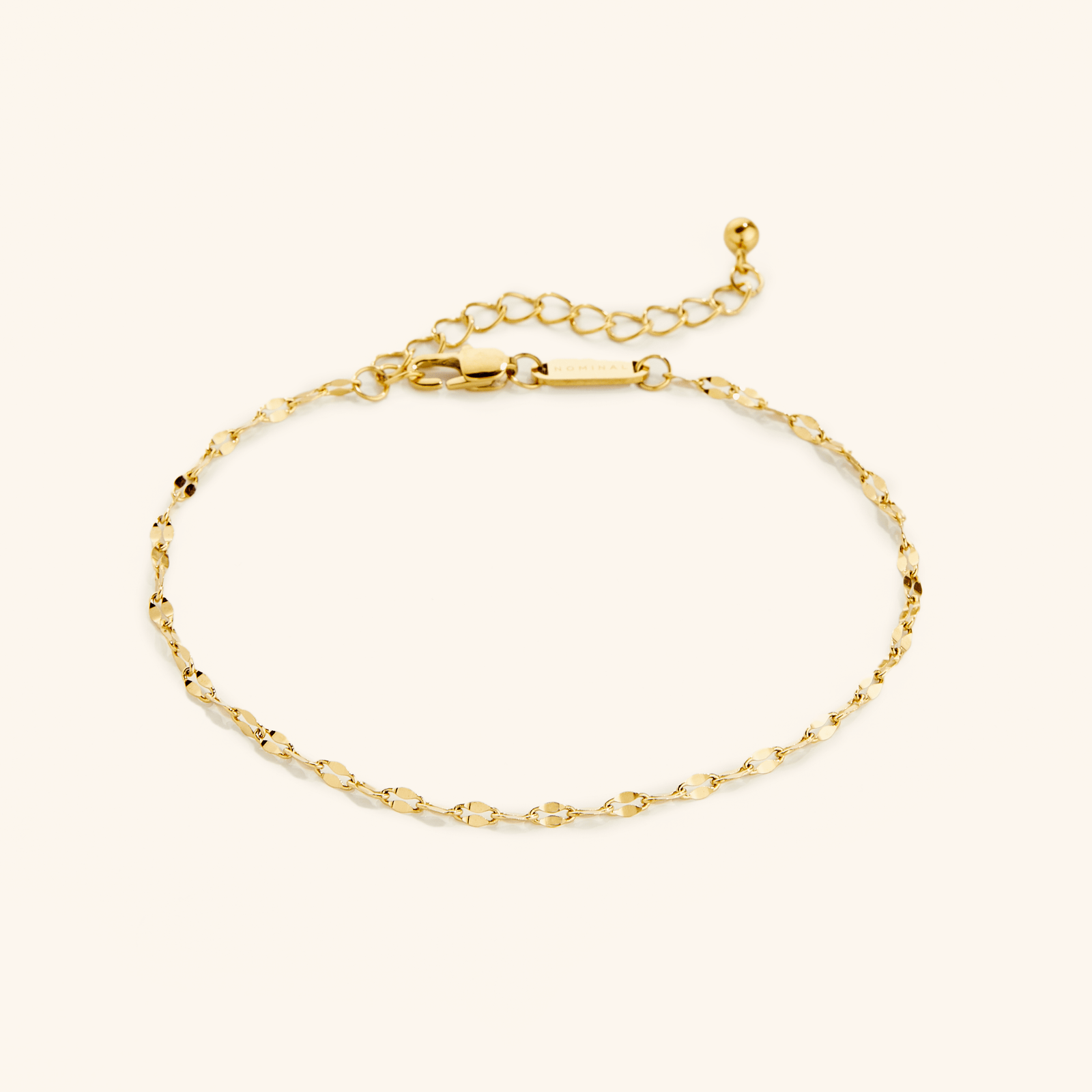 Flat lay of a delicate gold anklet with a flutter-style chain. The anklet features an adjustable extension chain with a small ball detail at the end and a clasp engraved with “NOMINAL”.