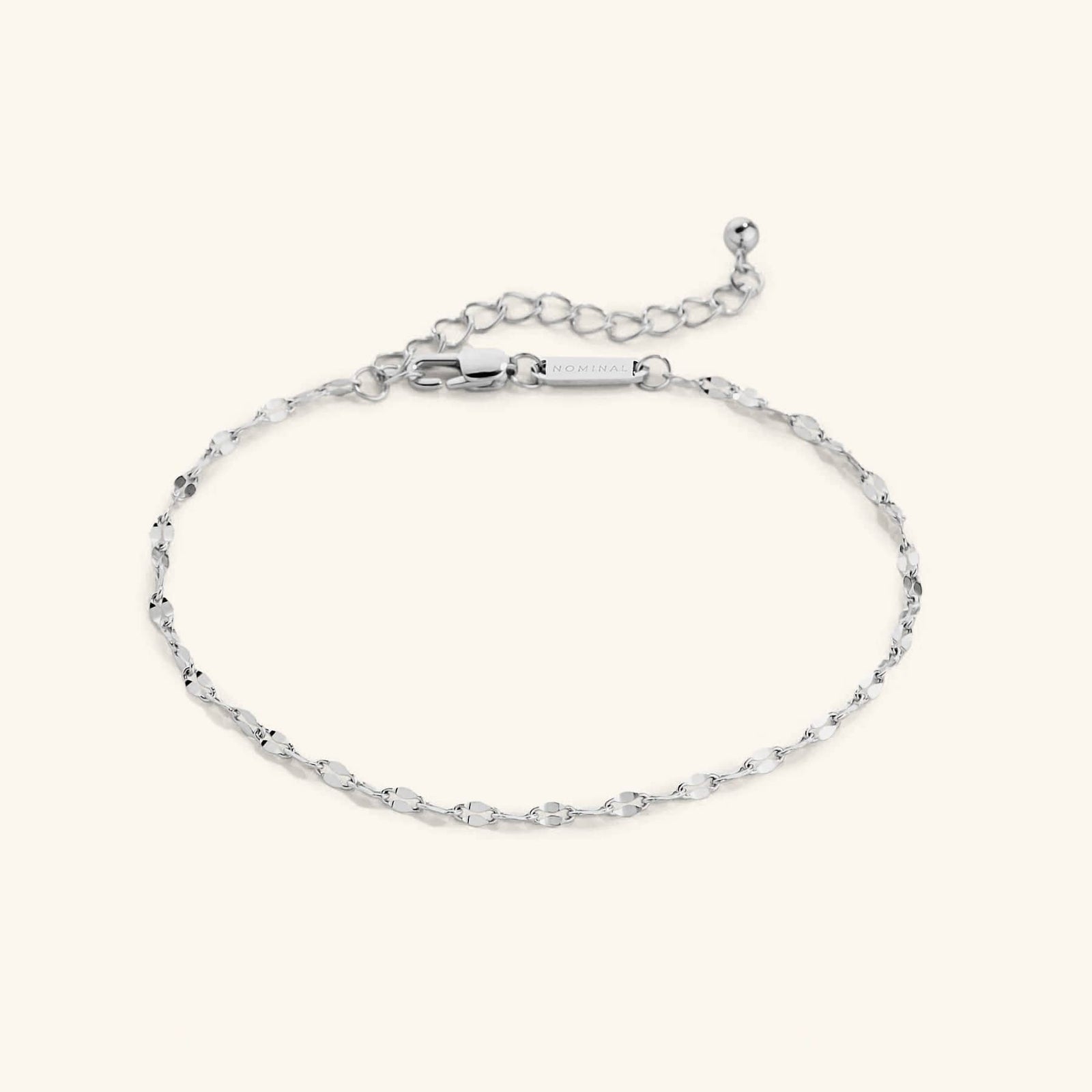 Flat lay of a delicate silver anklet with a flutter-style chain. The anklet features an adjustable extension chain with a small ball detail at the end and a clasp engraved with “NOMINAL”.