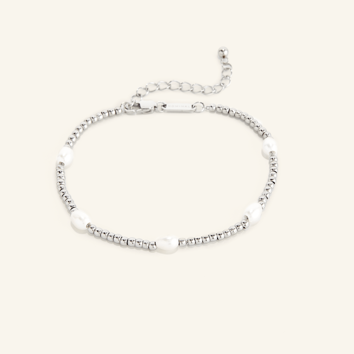 Silver anklet with small round beads and five evenly spaced white pearls, shown on a cream background with an adjustable clasp and extender chain engraved with 'NOMINAL'.