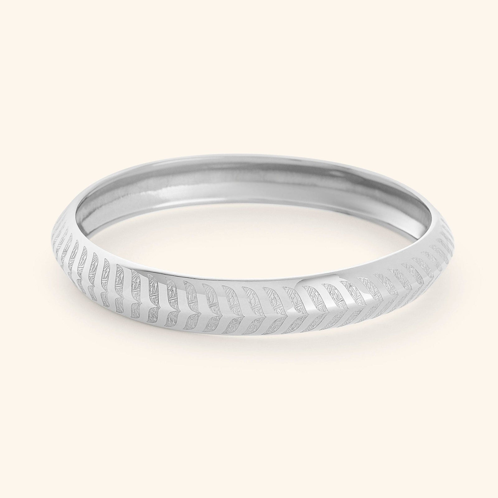 A silver "Chevron Bangle" designed for women as part of the Aadil x Nominal collection by Nominal. The bangle has a smooth, rounded surface and is decorated with intricate, stylized chevron or feather-like carvings that run around its entire circumference.