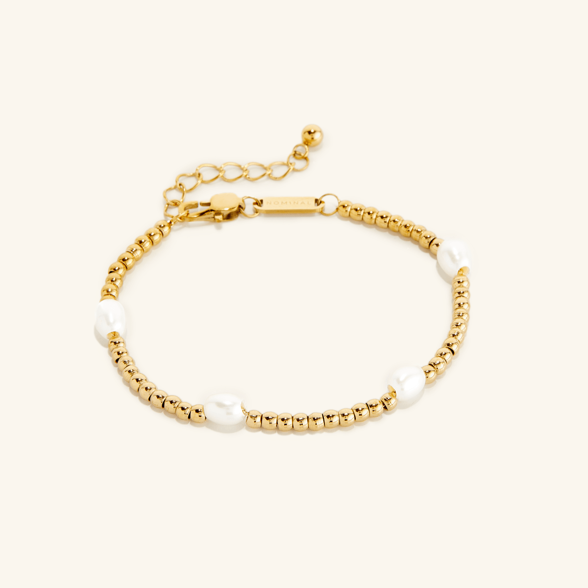 Gold bracelet with small round beads and five evenly spaced white pearls, shown on a cream background with an adjustable clasp and extender chain engraved with 'NOMINAL'.