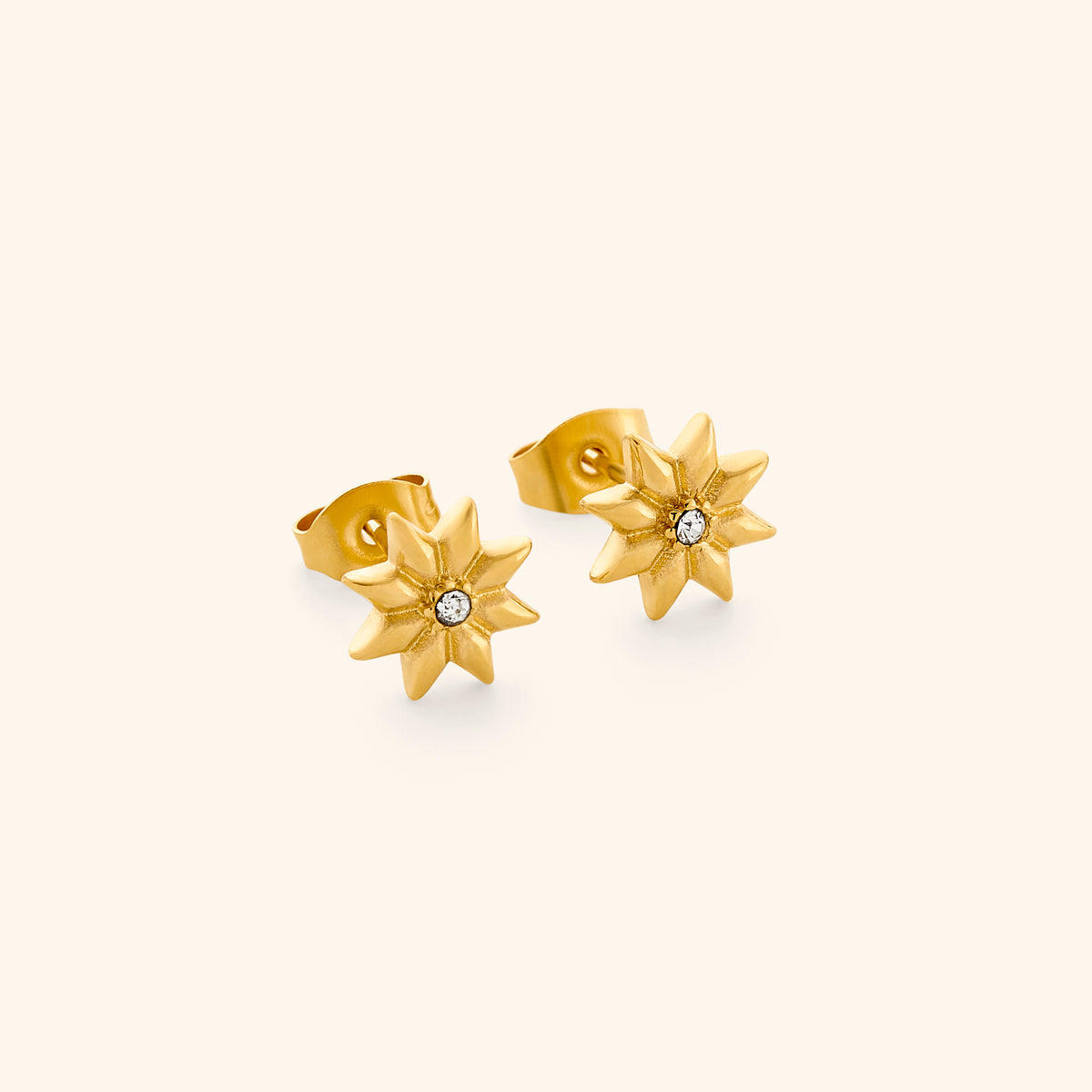 The Bezel Bloom Studs by Nominal, crafted with an 18k gold-plated finish featuring blooming starburst floral petals centered with a bezel-set crystal, displayed on an off-white background.
