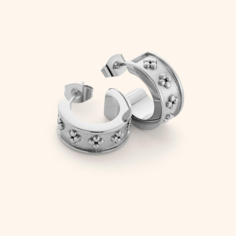 Flat lay of silver Henna Hoops pimage_ wide curved band with raised dotted floral motifs on a textured background.