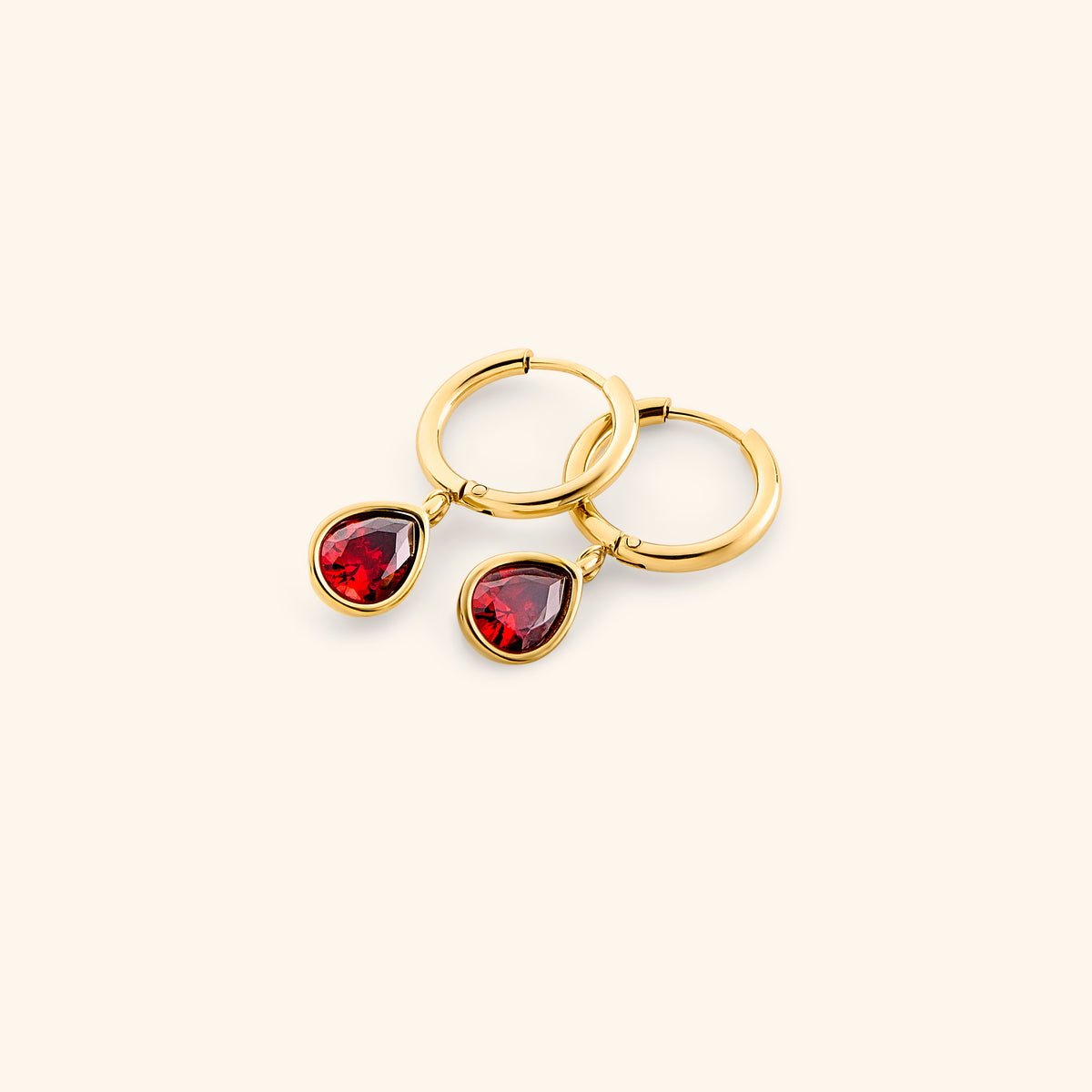 Alt text: The Garnet Stone Huggie Hoops by Nominal, crafted with an 18k gold-plated finish featuring smooth hoop bands accented with dangling teardrop red garnet gemstone charms, displayed on an off-white background.