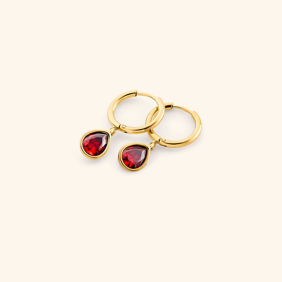 Alt text: The Garnet Stone Huggie Hoops by Nominal, crafted with an 18k gold-plated finish featuring smooth hoop bands accented with dangling teardrop red garnet gemstone charms, displayed on an off-white background.