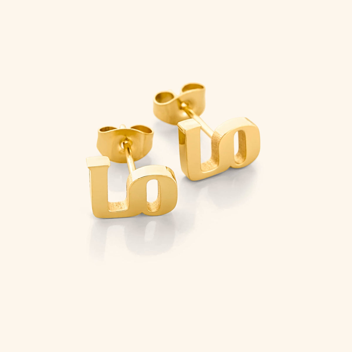 A close-up of the "Mama" stud earrings in 18K gold plating. Each earring features the Arabic letter "م" ("Meem"), and when worn together, they form the word "ماما" ("Mama"), symbolizing maternal love and strength. The gold finish adds a touch of warmth and elegance to the minimalist design.