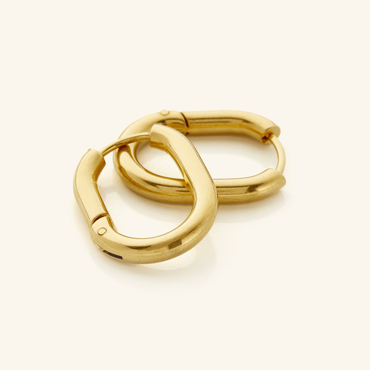 The image shows a pair of 18k gold plated hoop earrings placed on a clean, neutral background. The earrings have a smooth, polished surface that reflects light softly, giving them a warm, lustrous appearance. They are slightly oval in shape rather than perfectly circular, which adds a subtle design detail.