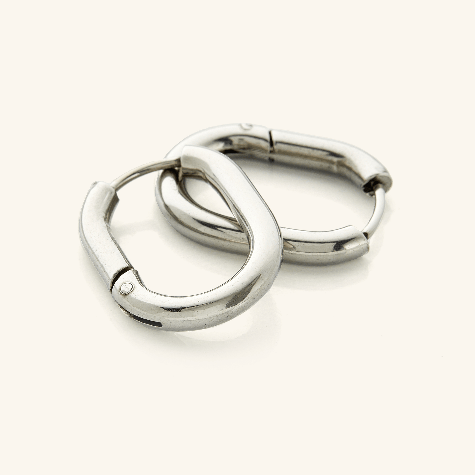 The image shows a pair of silver hoop earrings placed on a clean, neutral background. The earrings have a smooth, polished surface that reflects light softly, giving them a warm, lustrous appearance. They are slightly oval in shape rather than perfectly circular, which adds a subtle design detail.