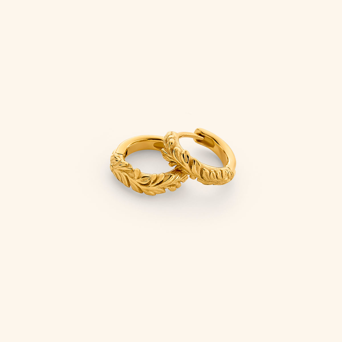 The Palestinian Olive Huggie Hoops by Nominal, featuring intricate embossed olive branch leaf detailing on 18k gold-plated hoop earrings, displayed on an off-white background.