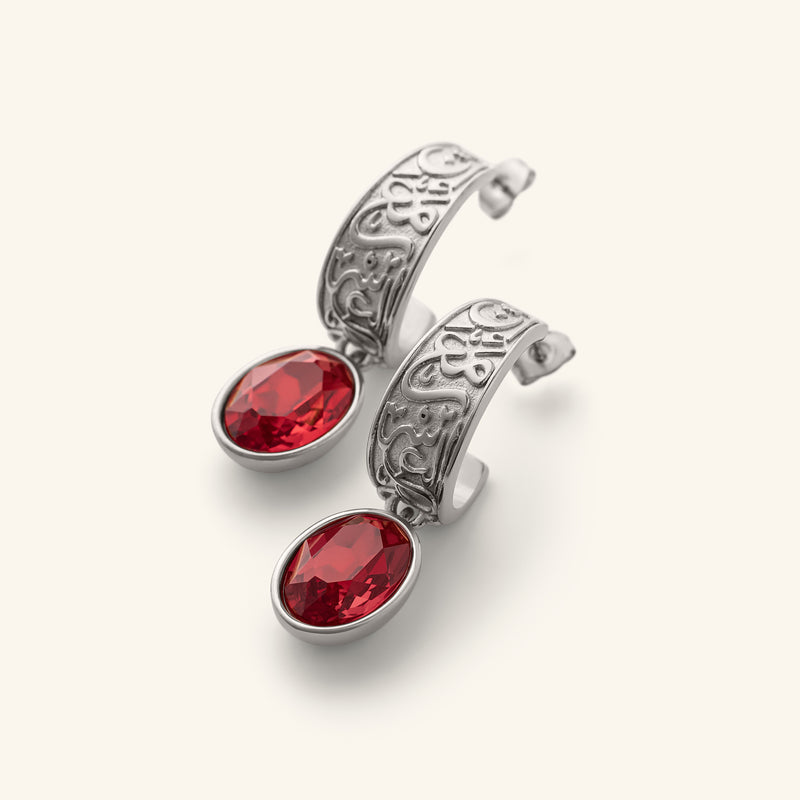 "With Hardship Comes Ease” Earrings in silver pimage_ featuring engraved Arabic calligraphy along the curved band with an oval red gemstone drop hanging below pimage_ styled against a neutral background.