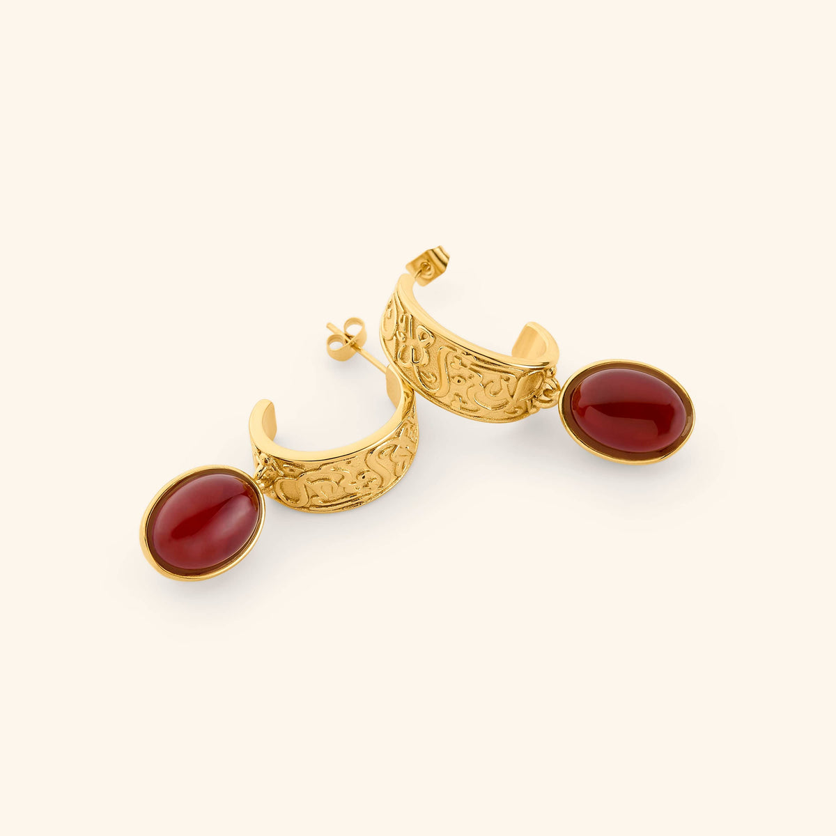 A pair of 18k gold plated hoop earrings against a light background. The earrings feature a vintage-style molded pair of hoops that read the most comforting promise in the Qur'an: "with every hardship comes ease". Finished with a red gem hanging from the hoops.
