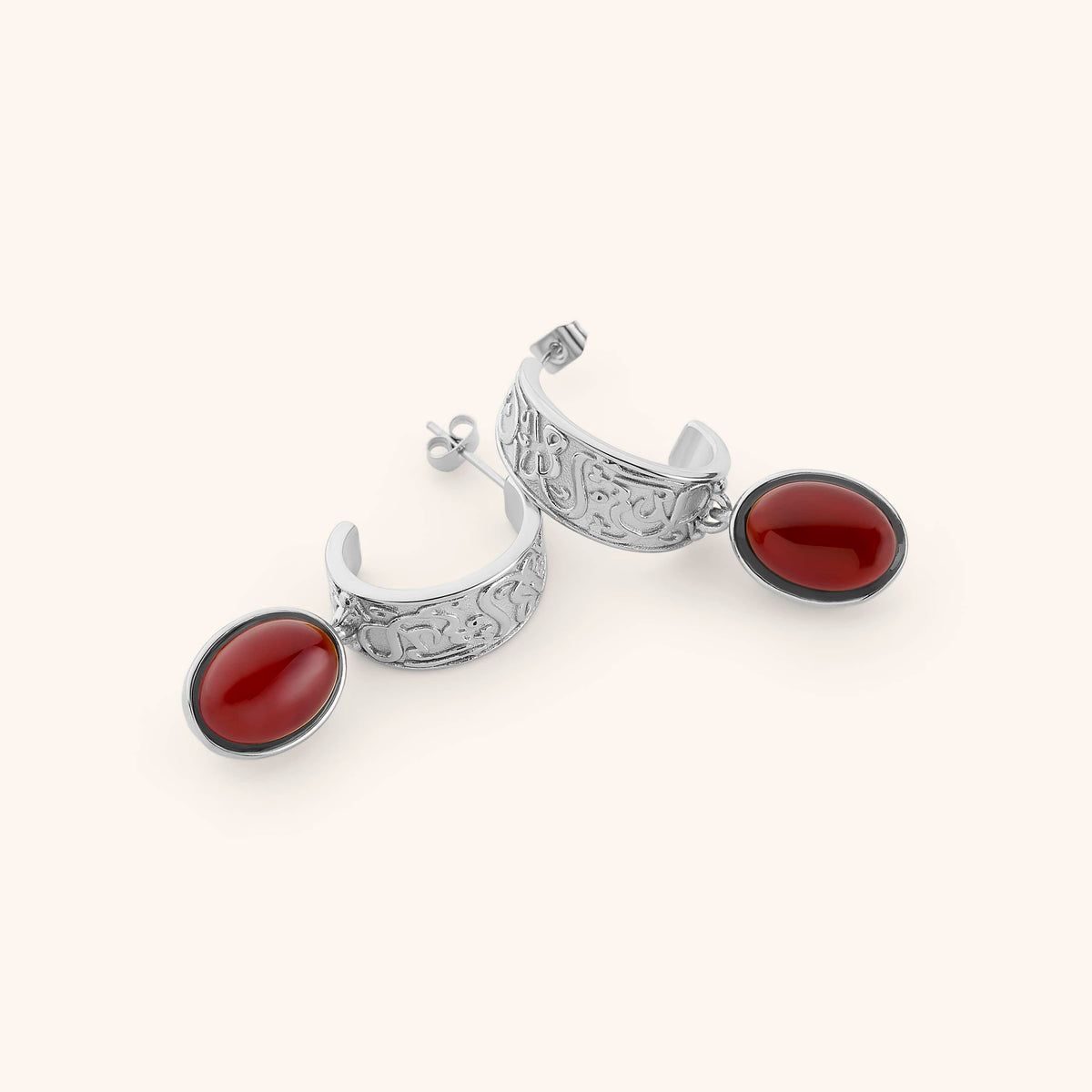 A pair of silver hoop earrings against a light background. The earrings feature a vintage-style molded pair of hoops that read the most comforting promise in the Qur'an: "with every hardship comes ease". Finished with a red gem hanging from the hoops.