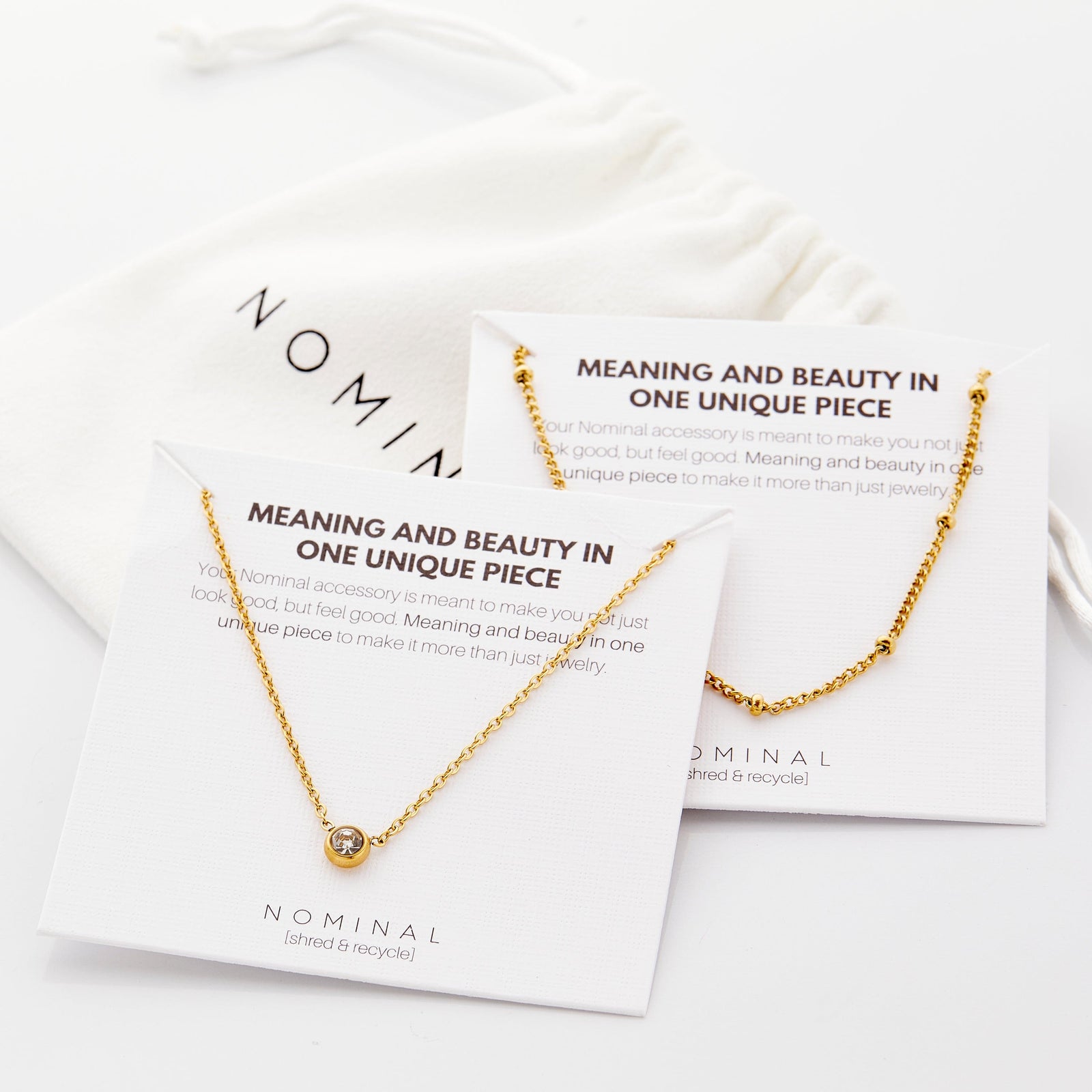 Necklace set featuring a bead-accented chain layered with a solitaire bezel-set pendant necklace displayed on branded Nominal cards with a white pouch. Card text (both cards):
“MEANING AND BEAUTY IN ONE UNIQUE PIECE
Your Nominal accessory is meant to make you not just look good, but feel good. Meaning and beauty in one unique piece to make it more than just jewelry.”
Brand text: NOMINAL [shred & recycle]