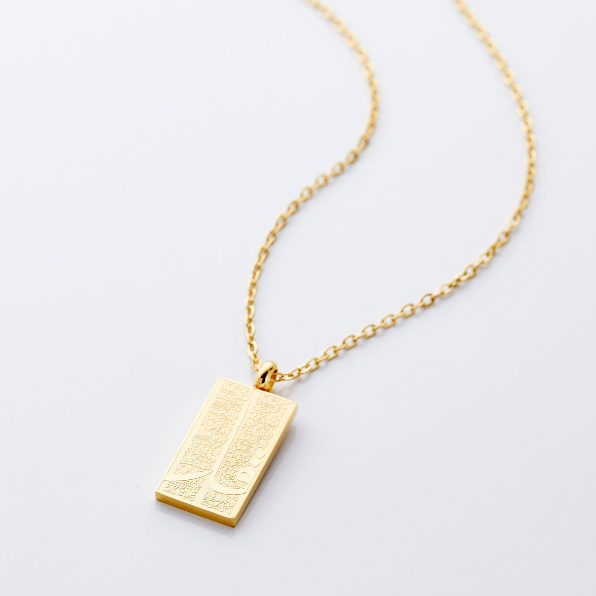 The image shows a delicate gold necklace displayed against a clean, light gray background. The chain is thin and elegant, forming a gentle curve that draws the eye toward the pendant hanging at the center.

The pendant is rectangular and made of gold, with a smooth, polished finish that reflects light subtly. Its surface features intricate engraved patterns that add texture and visual interest.