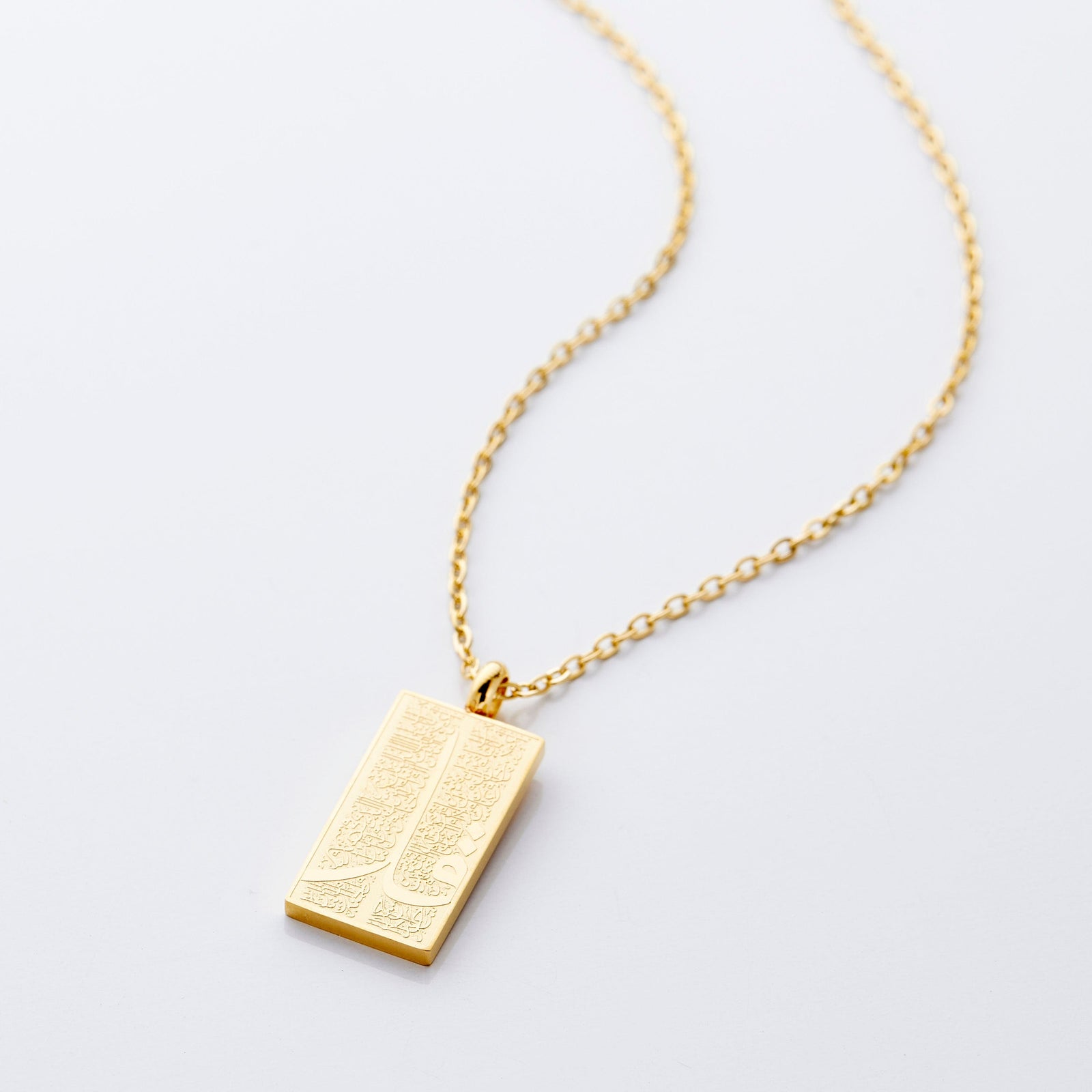 The image shows a delicate gold necklace displayed against a clean, light gray background. The chain is thin and elegant, forming a gentle curve that draws the eye toward the pendant hanging at the center.

The pendant is rectangular and made of gold, with a smooth, polished finish that reflects light subtly. Its surface features intricate engraved patterns that add texture and visual interest.