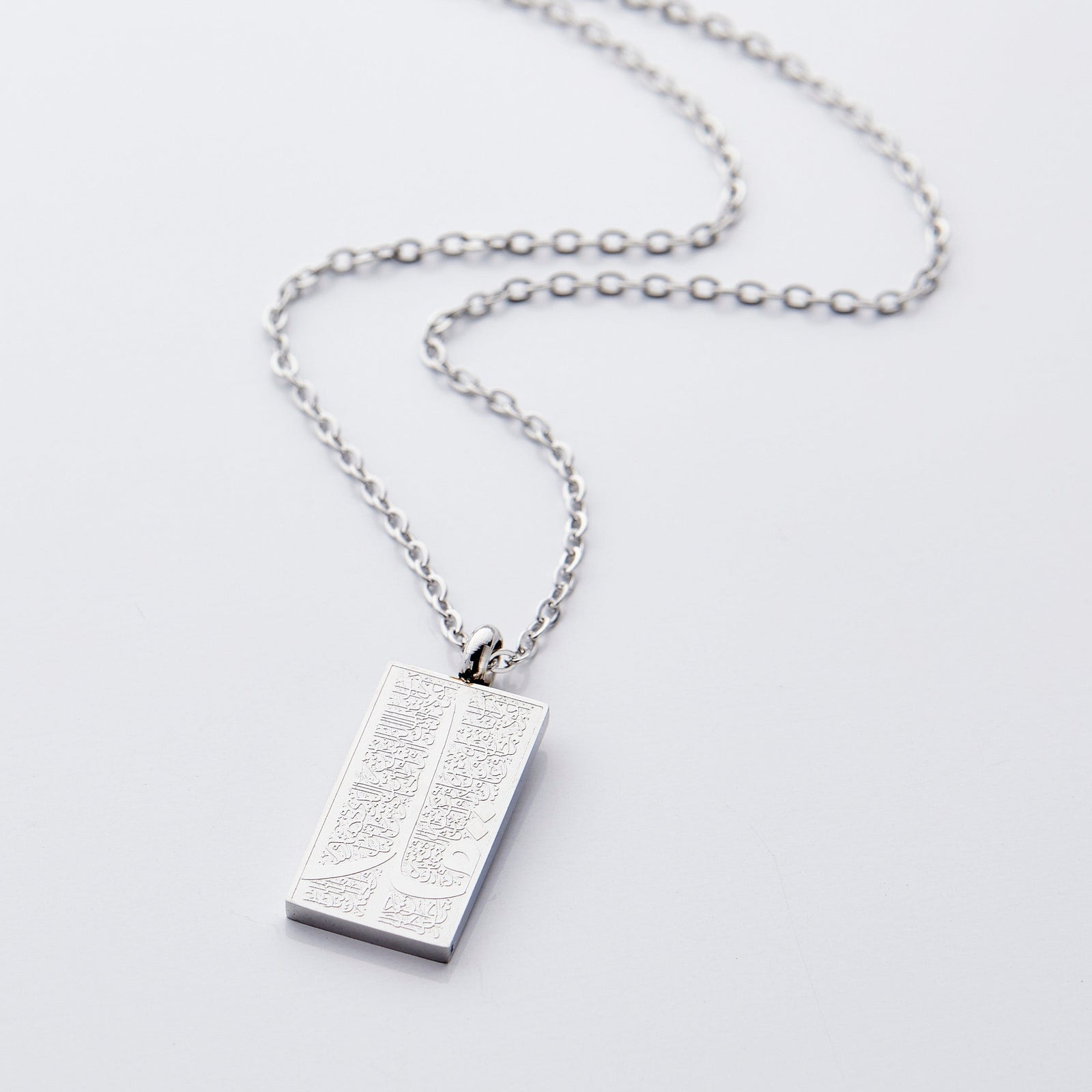 The image shows a delicate gold necklace displayed against a clean, light gray background. The chain is thin and elegant, forming a gentle curve that draws the eye toward the pendant hanging at the center.

The pendant is rectangular and made of silver, with a smooth, polished finish that reflects light subtly. Its surface features intricate engraved patterns that add texture and visual interest.