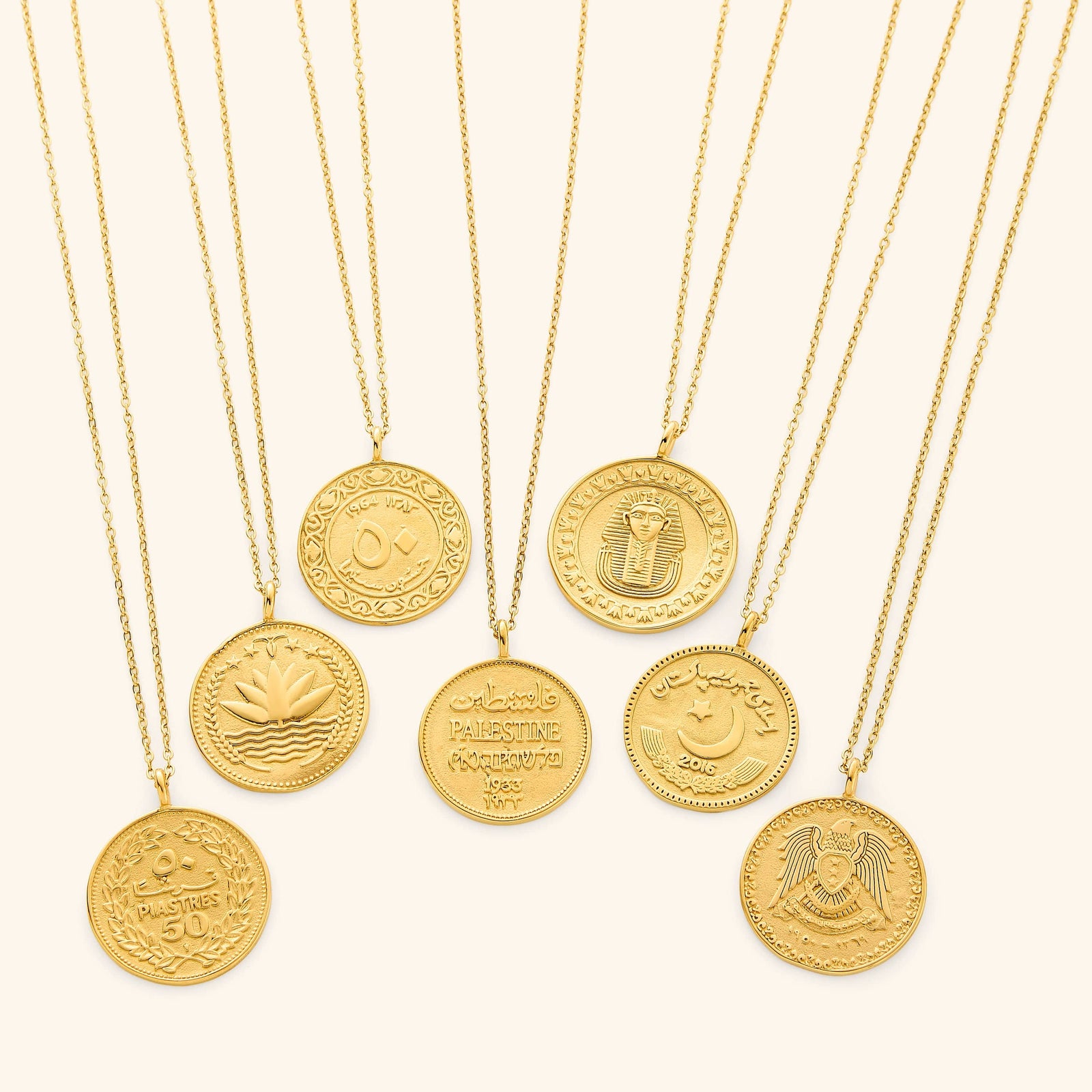 Seven gold-plated country coin necklaces arranged side by side on a neutral background. Each pendant showcases a unique engraved coin design representing a different country, featuring intricate Arabic script, national emblems, and cultural symbols. The 18K gold finish unifies the collection while highlighting the distinct details of each coin.