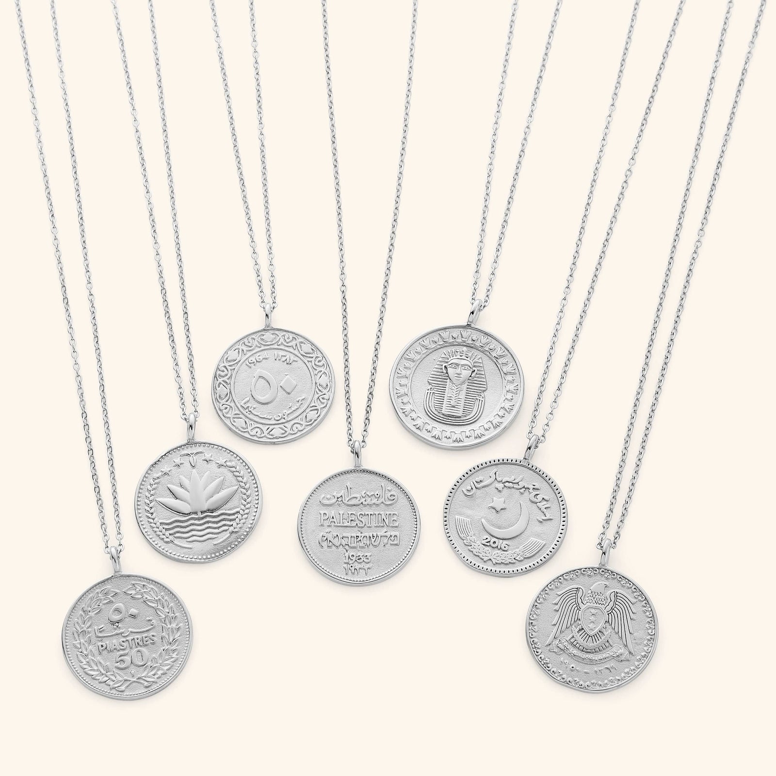Seven silver country coin necklaces displayed in a row on a neutral backdrop. Each round pendant features a unique engraved design representing a different country’s coin, with intricate Arabic script, national symbols, and cultural motifs. The polished silver finish gives the collection a cohesive and elegant appearance while showcasing the individuality of each coin.