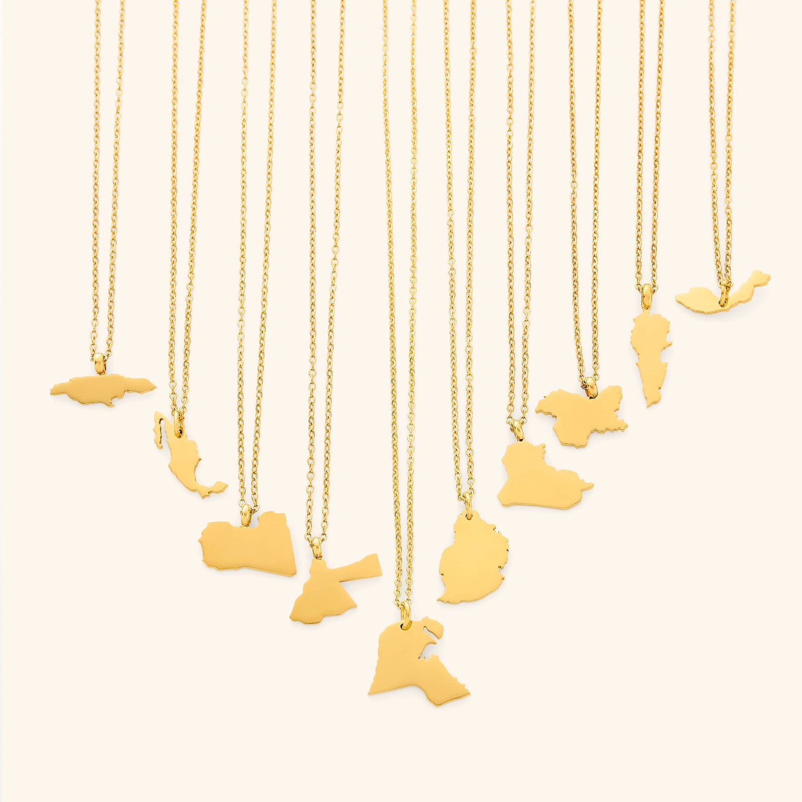 The image shows a collection of delicate gold necklaces hanging vertically against a light beige background. Each necklace features a flat, polished gold pendant shaped like the outline of a different country or region.