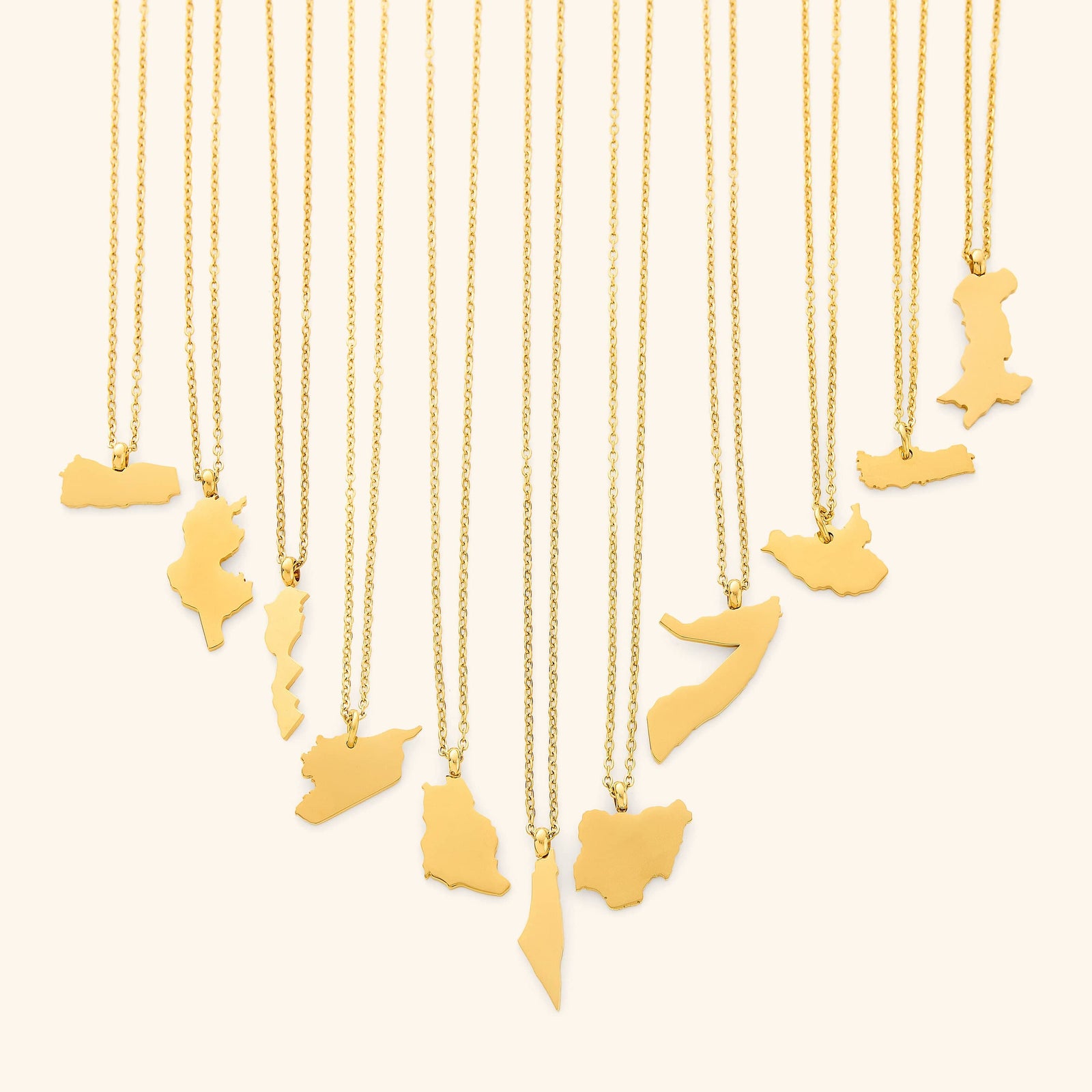 The image shows a collection of delicate gold necklaces hanging vertically against a light beige background. Each necklace features a flat, polished gold pendant shaped like the outline of a different country or region.