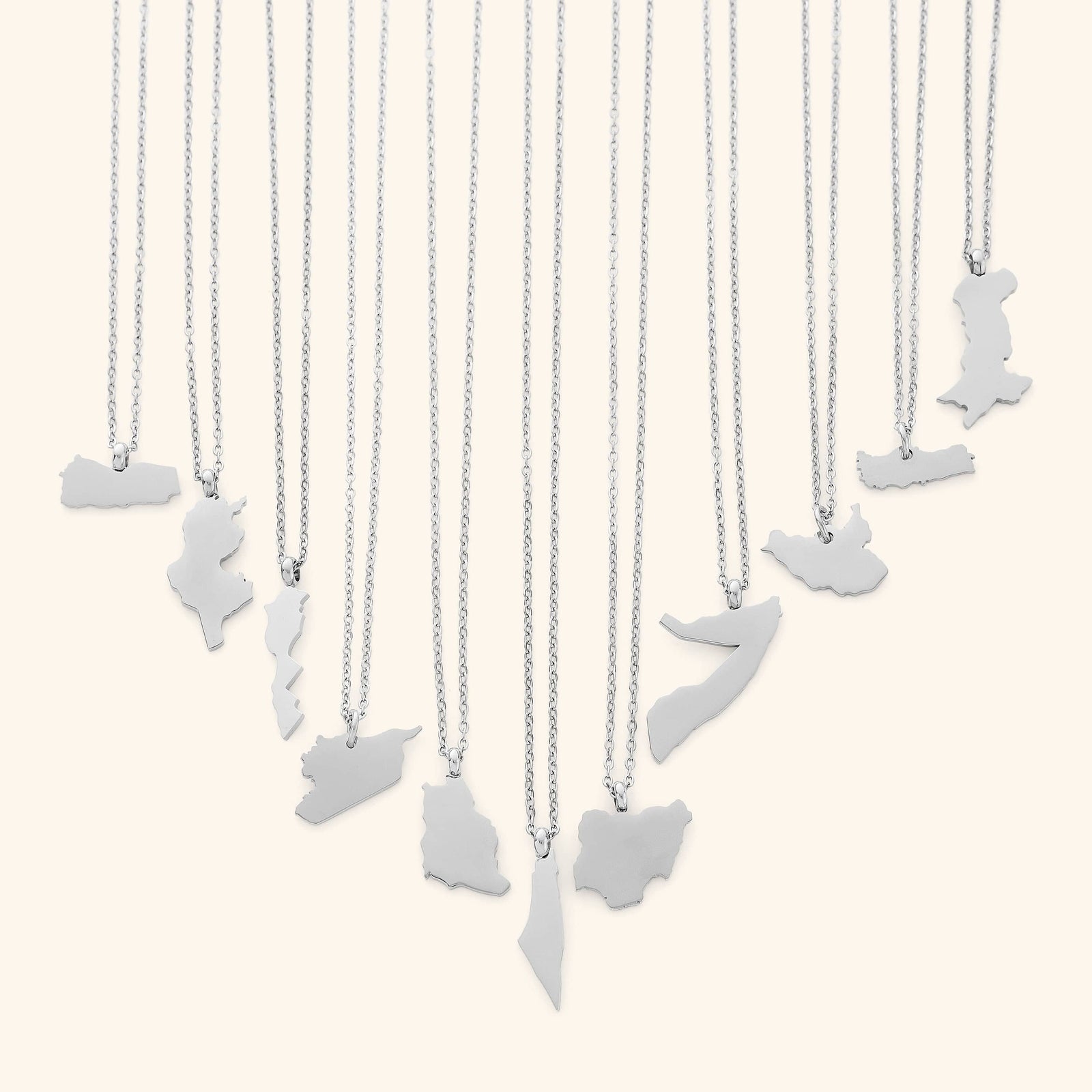 The image shows a collection of delicate silver necklaces hanging vertically against a light beige background. Each necklace features a flat, polished silver pendant shaped like the outline of a different country or region.