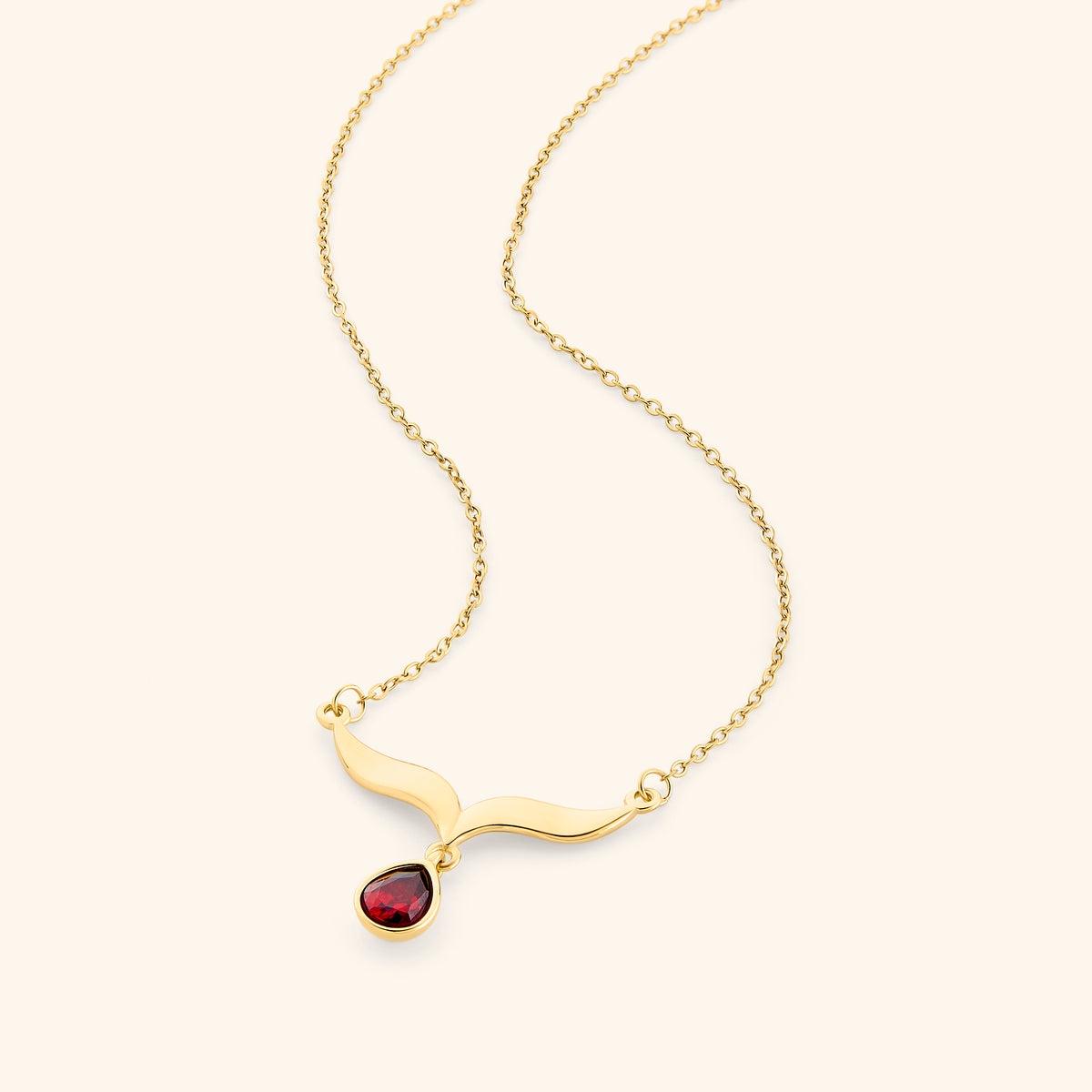 The Garnet Stone Keffiyeh Necklace by Nominal, crafted with an 18k gold-plated finish featuring a sculpted curved keffiyeh pendant suspending a teardrop red garnet gemstone, displayed on an off-white background.
