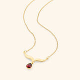 The Garnet Stone Keffiyeh Necklace by Nominal, crafted with an 18k gold-plated finish featuring a sculpted curved keffiyeh pendant suspending a teardrop red garnet gemstone, displayed on an off-white background.