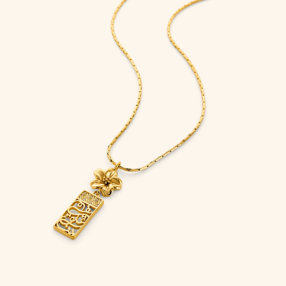 18K Gold plated necklace with rectangular pendant that features patterns found in the Damascus masjids and courtyards, bordering the Arabic calligraphy for "in our hearts" topped with a blooming Jasmine flower on a white background.