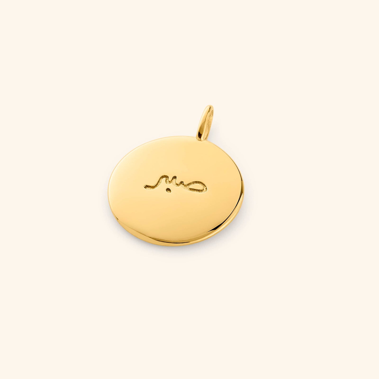 Gold round charm engraved with the Arabic word "Sabr", meaning patience, placed flat on a soft beige background.