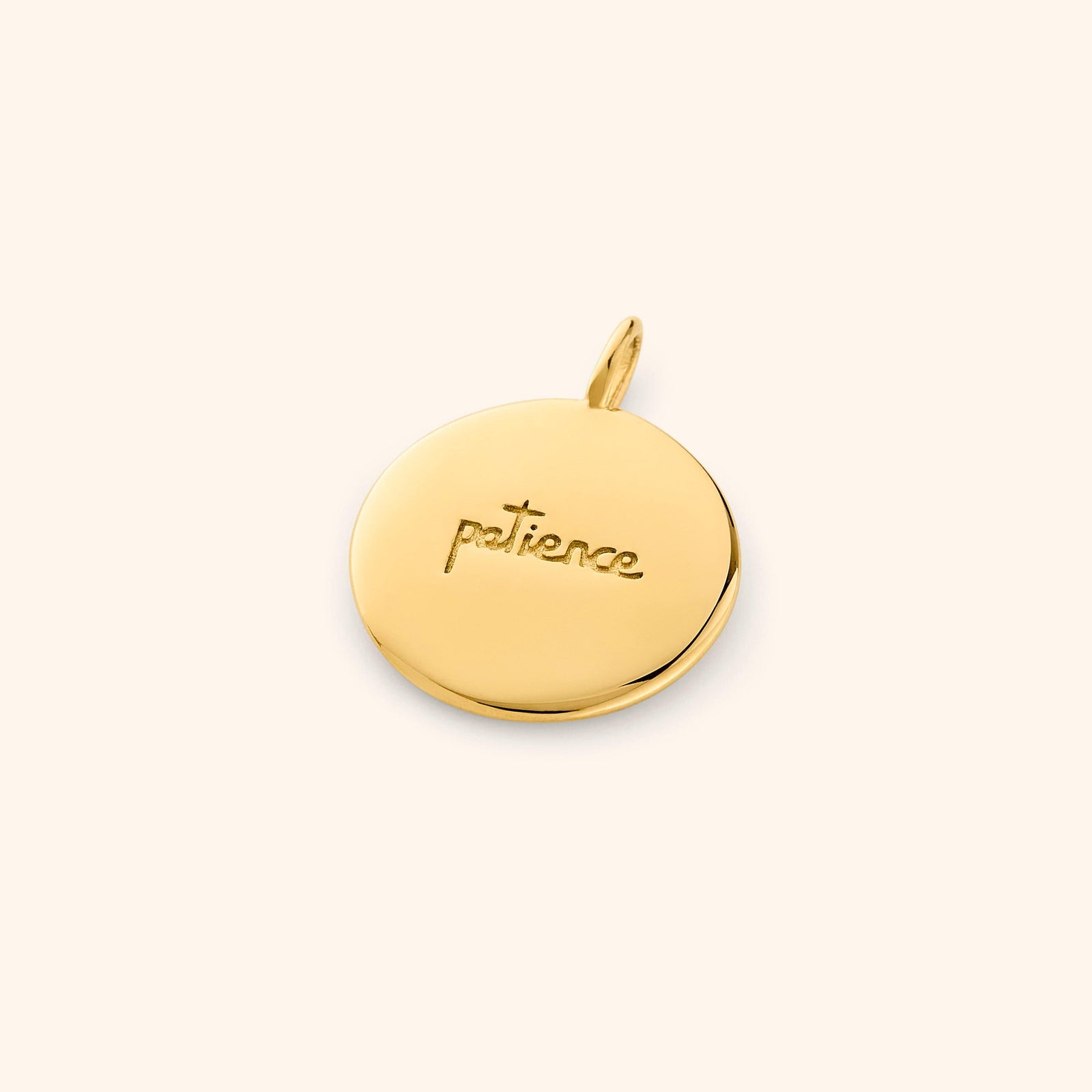 Reverse side of the gold charm engraved with the word "patience", resting flat on a beige backdrop.