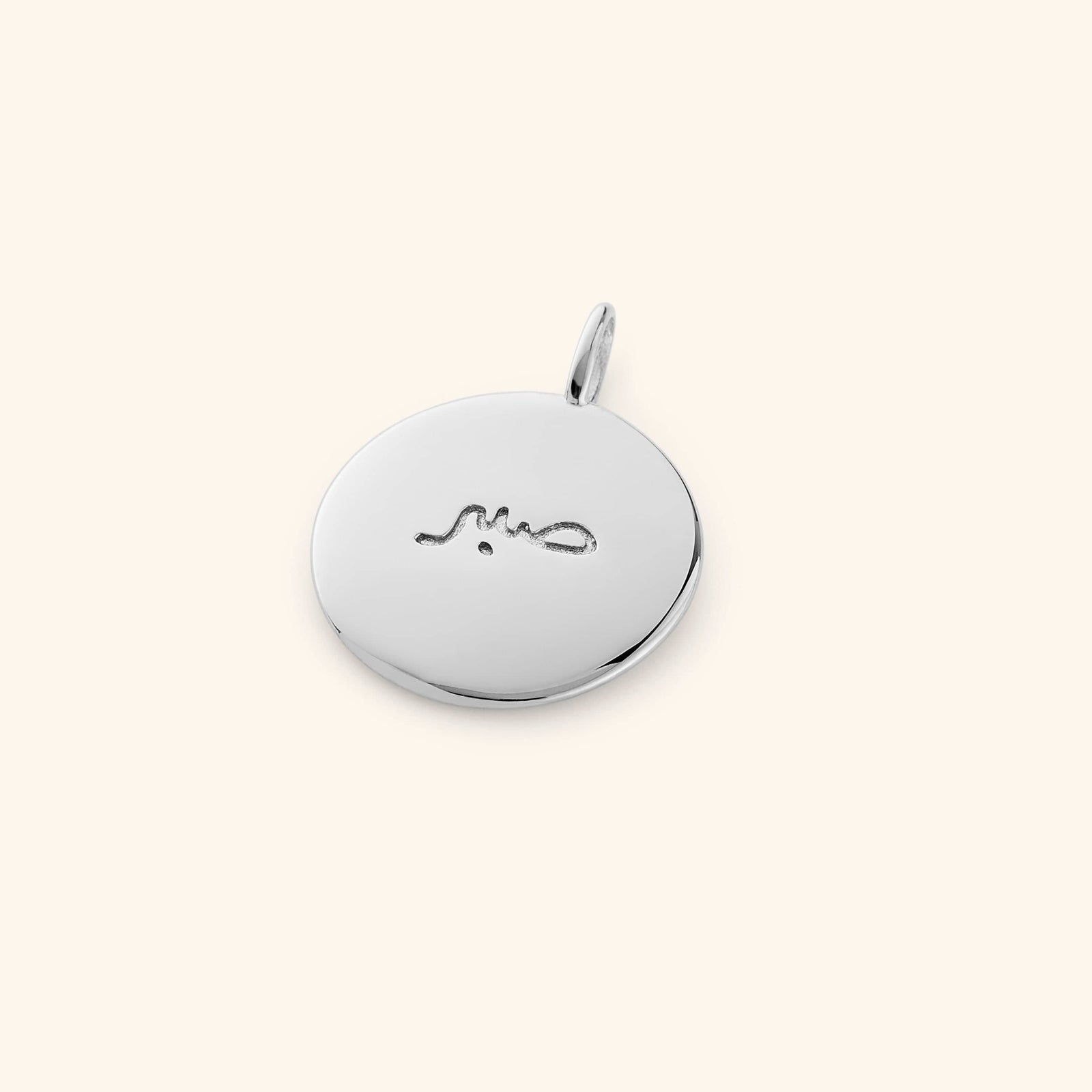 Silver round charm engraved with the Arabic word "Sabr", lying flat on a beige background.