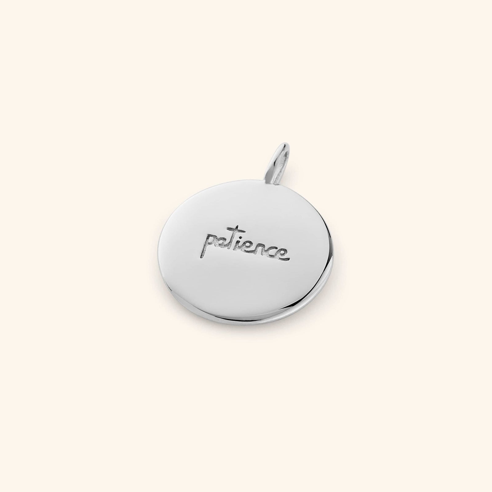 Silver Sabr charm showing the word "patience" engraved in English, laid flat on a soft beige surface.