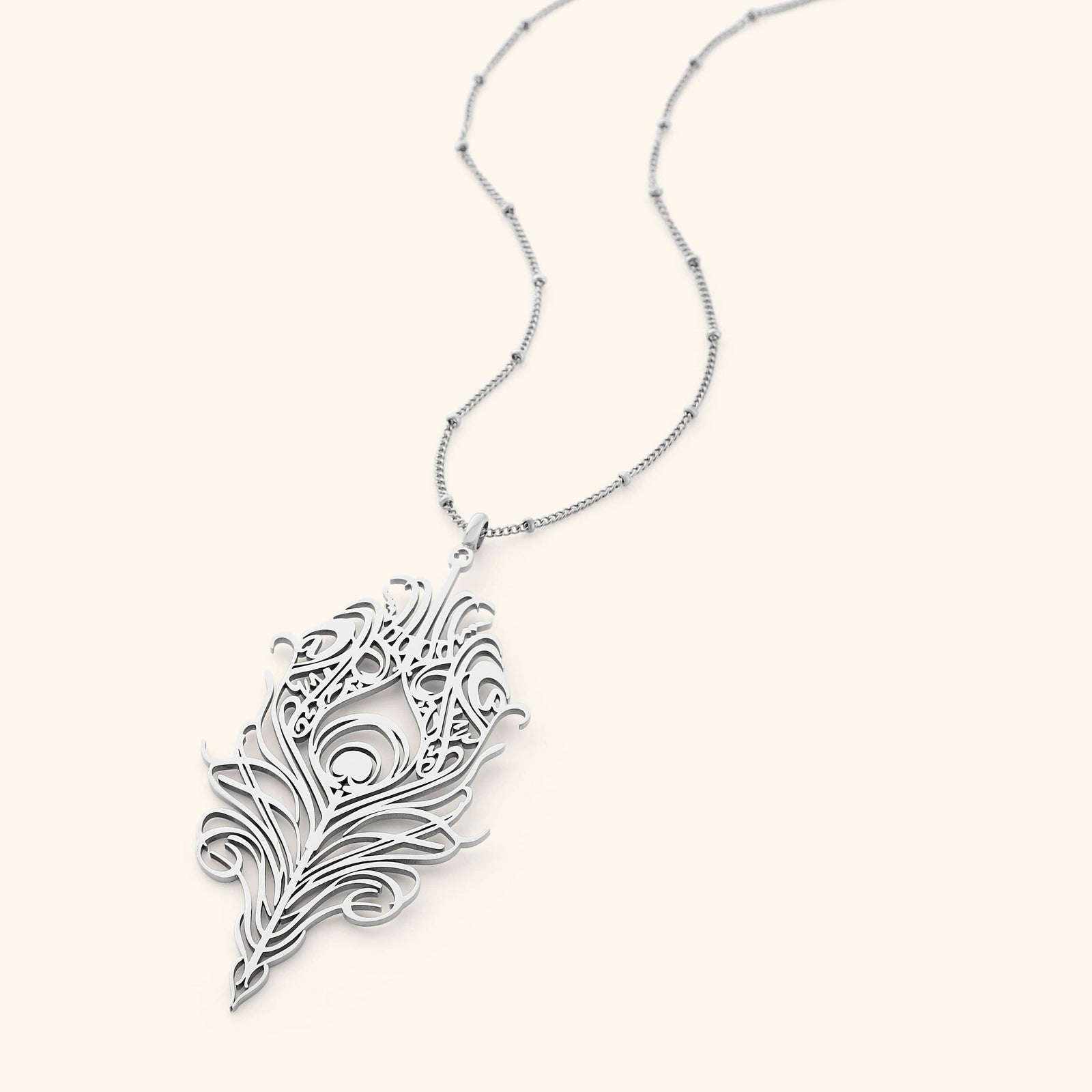 A delicate silver-plated chain necklace featuring a stylized, peacock feather-shaped pendant with intricate cut-out details. The pendant and chain are displayed against a light beige background.