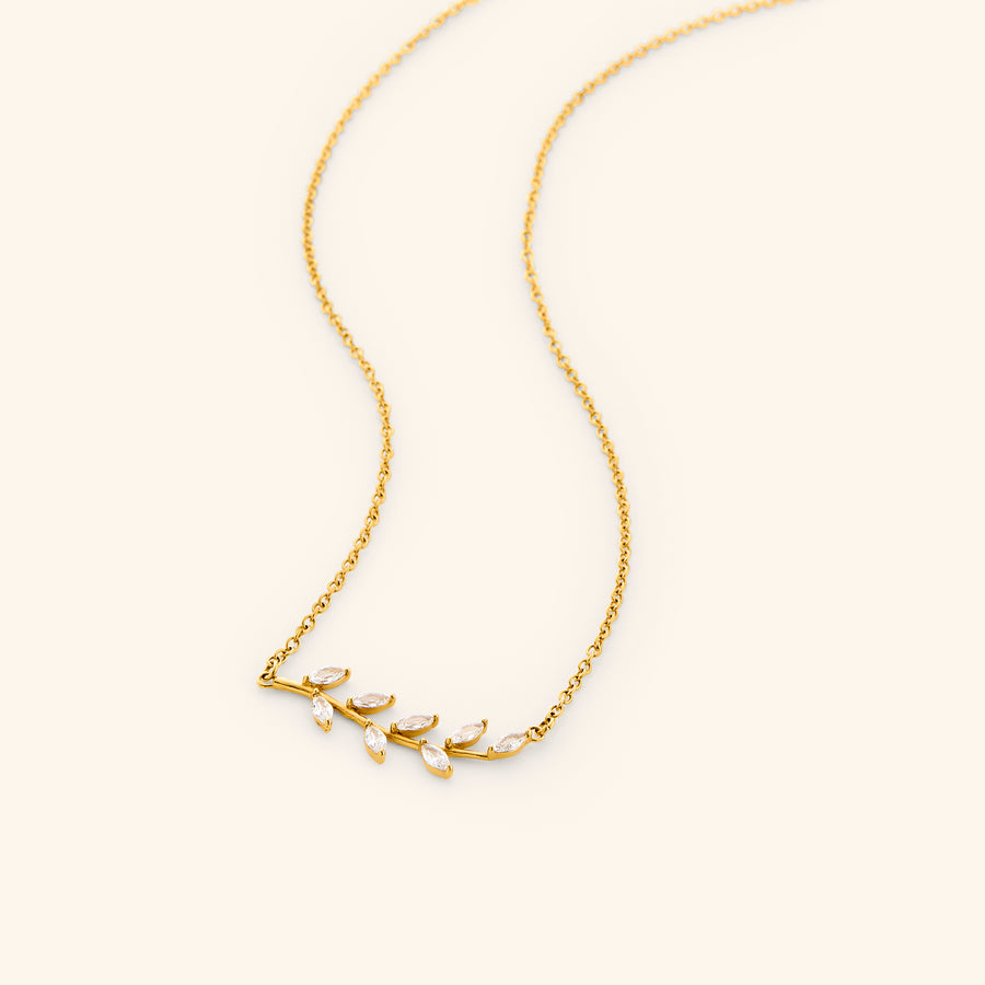 The Pavé Olive Branch Necklace by Nominal, featuring a delicate 18k gold-plated chain with a curved horizontal olive branch pendant set with sparkling marquise crystals, displayed on an off-white background.