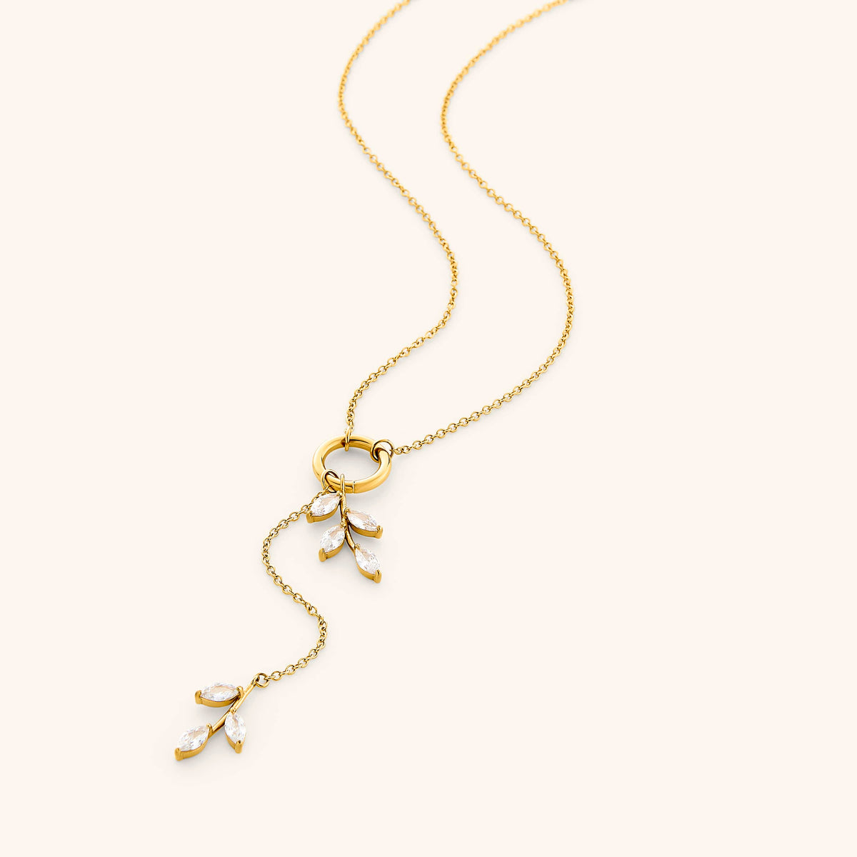 The Pavé Olive Branch Lariat Necklace by Nominal, featuring a delicate 18k gold-plated chain joined by a central ring with asymmetrical sparkling crystal olive leaf drops, displayed on an off-white background.