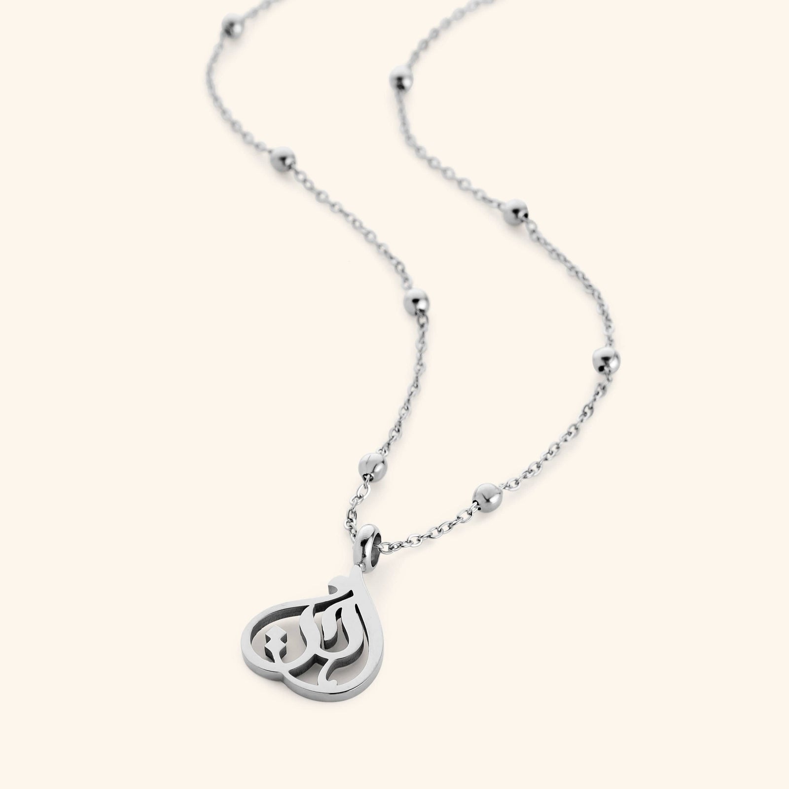 Silver necklace with beaded chain and teardrop-shaped pendant featuring Arabic calligraphy spelling “Sister”, photographed flat against a cream background.