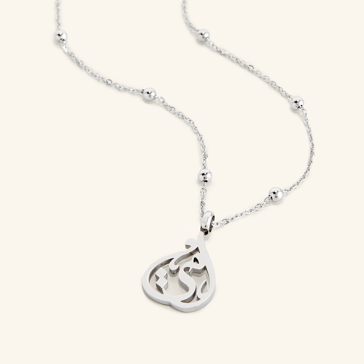 Flat lay of a silver necklace that features a gorgeous cradled shape featuring an Arabic calligraphy of "ummi" / "my mother".