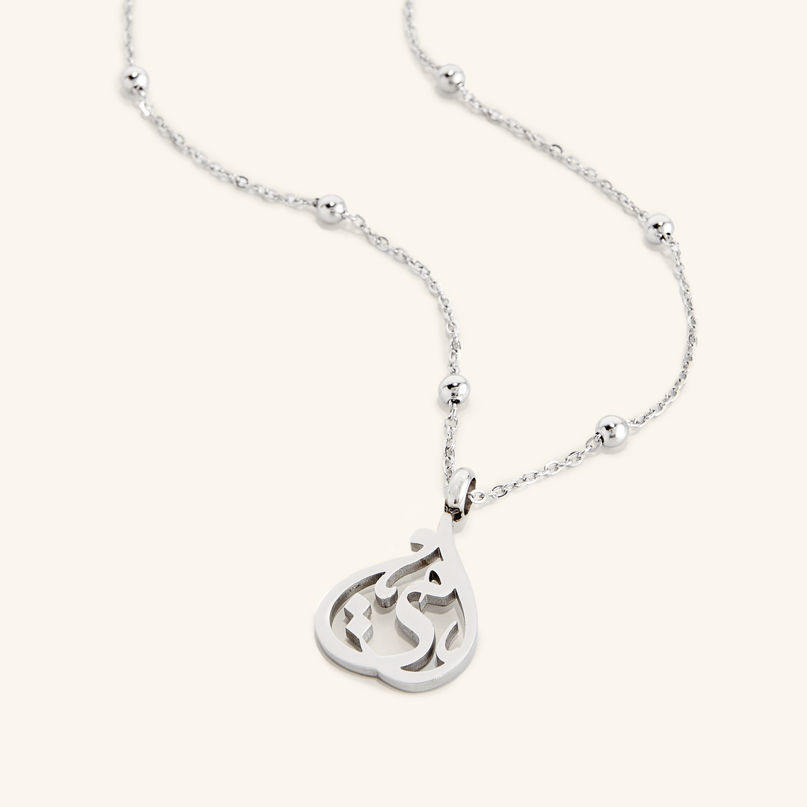 Flat lay of a silver necklace that features a gorgeous cradled shape featuring an Arabic calligraphy of "ummi" / "my mother".