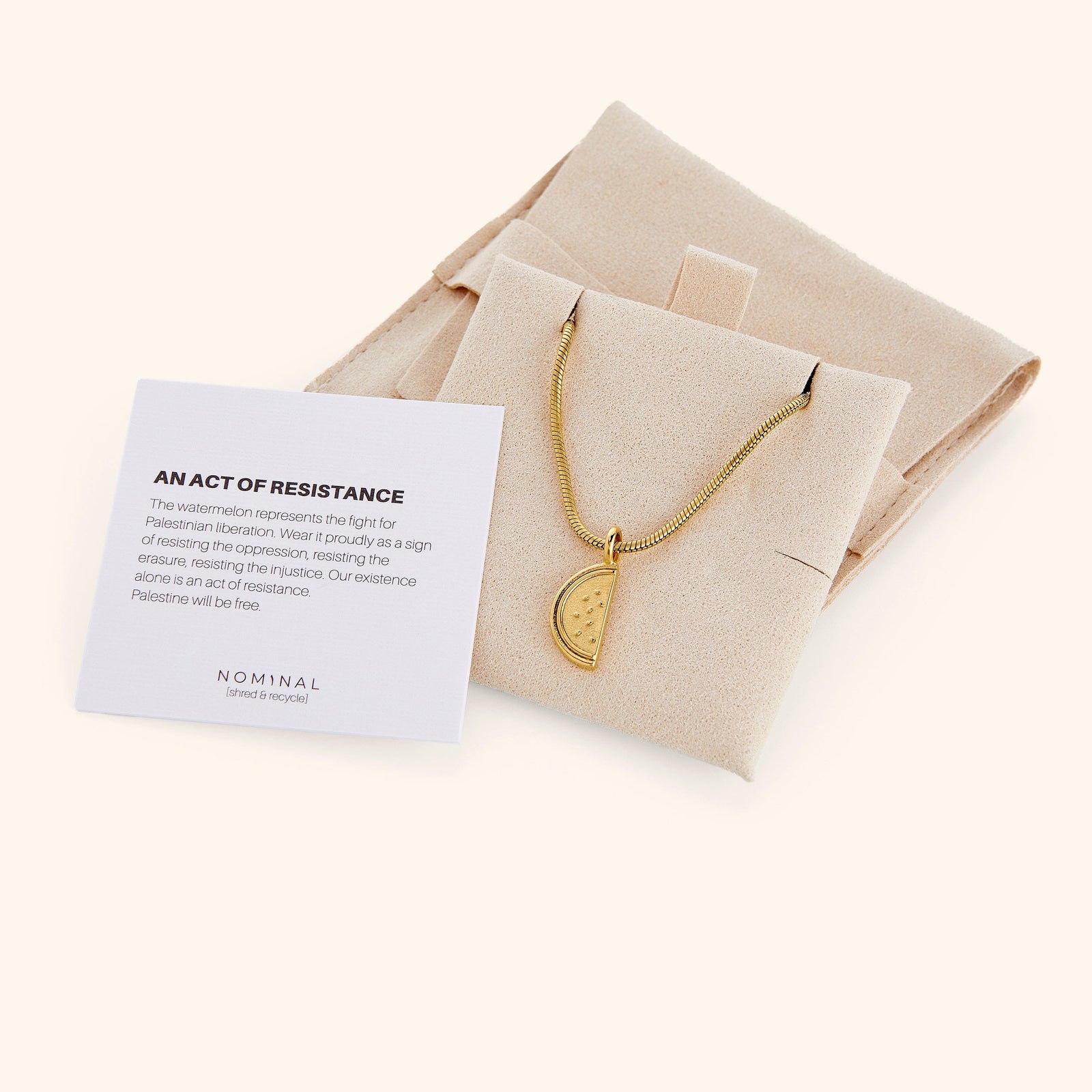 A gold necklace with a watermelon slice pendant is nestled in a light beige pouch. Next to the pouch is a small white card with black text that reads "AN ACT OF RESISTANCE" at the top, followed by a paragraph of smaller text and the word "NOMINAL" with "a jewlery brand" underneath at the bottom left. The pouch and card are sitting on a light beige surface.
