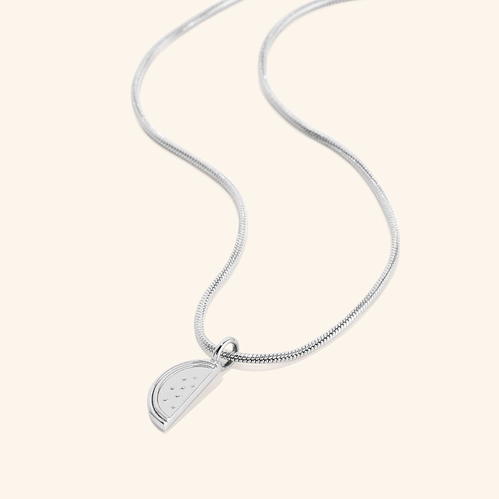 A silver necklace with a pendant shaped like a slice of a watermelon. The watermelon pendant has small indentations representing seeds. The necklace features a thin, flexible silver snake chain. The necklace is displayed on a light beige background.