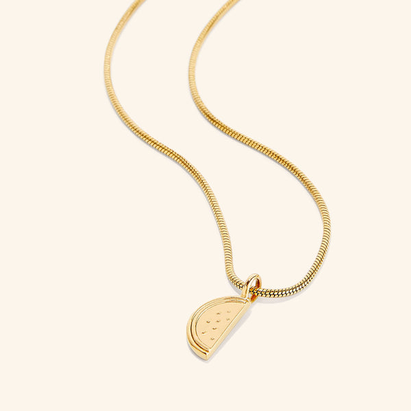 A gold necklace with a pendant shaped like a slice of a watermelon. The watermelon pendant is detailed with small indentations representing seeds. The necklace chain is a thin, flexible gold snake chain. The necklace is displayed against a light beige background.